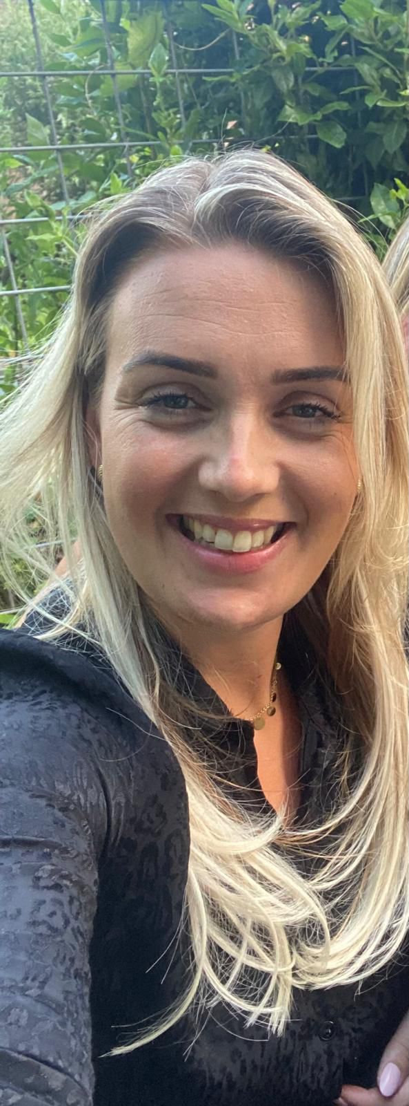 Close-up of a smiling woman with long blonde hair wearing a black patterned top and a gold necklace, outdoors with greenery in the background.
