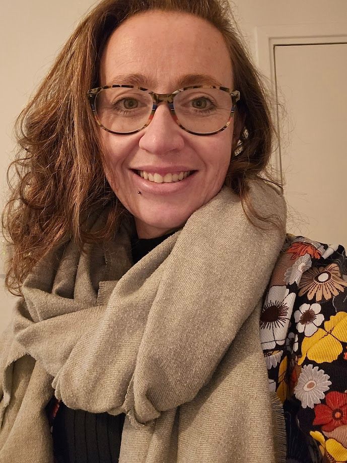 Smiling woman with curly brown hair wearing tortoiseshell glasses, a beige scarf, and a floral patterned top.