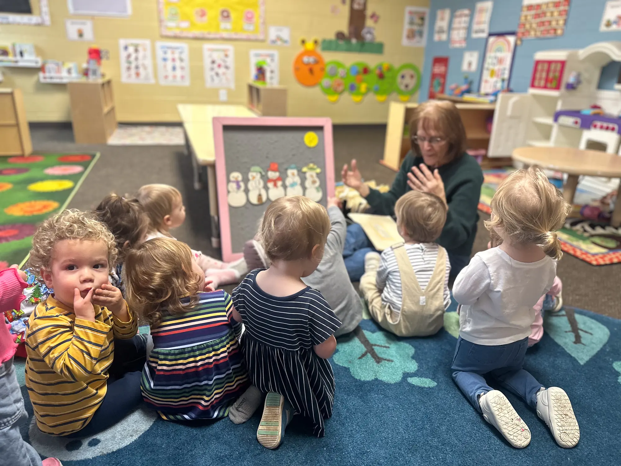 Toddlers sitting on a classroom rug while a teacher leads a felt-board story