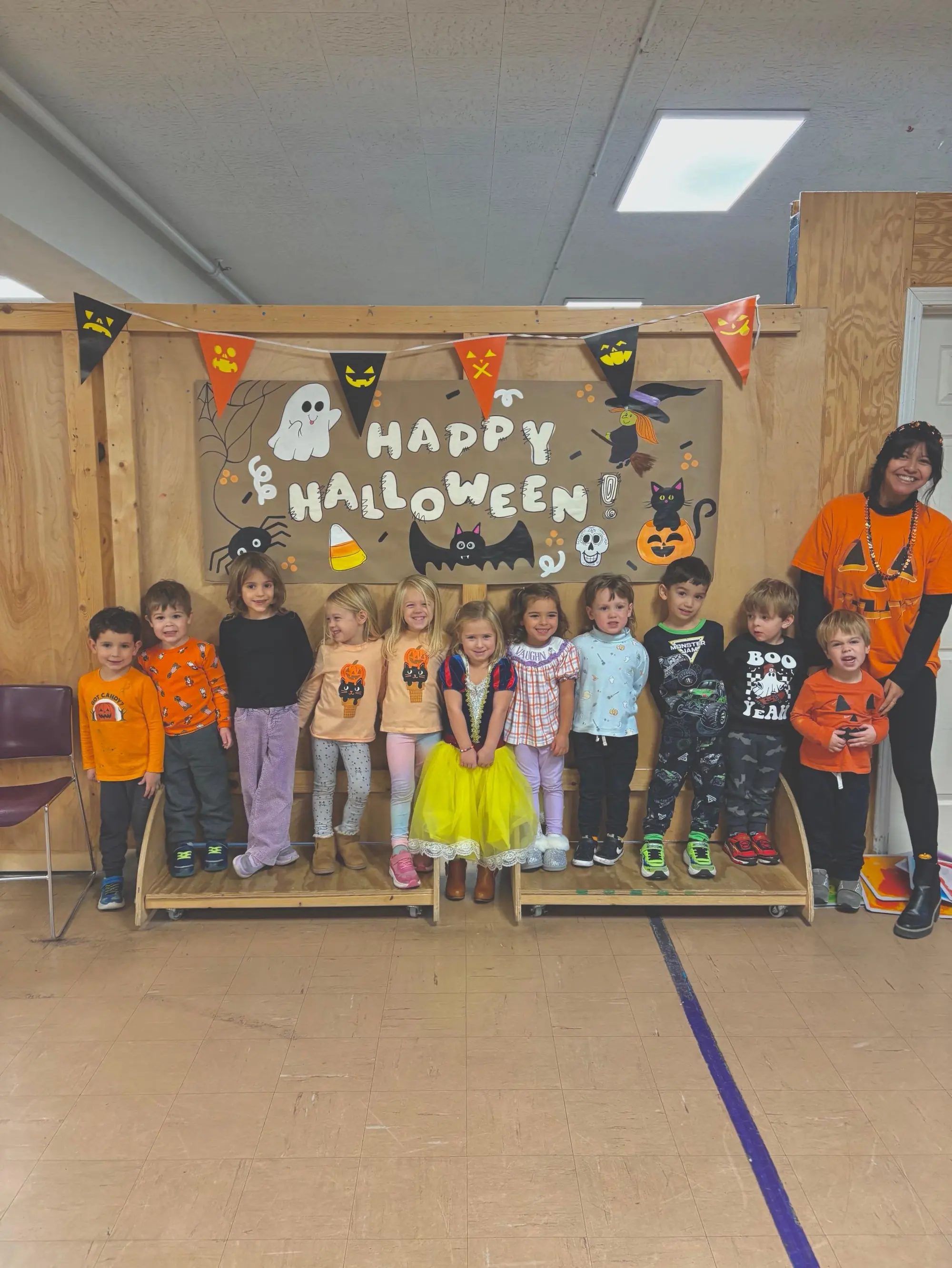 A class of preschoolers in Halloween costumes lined up with their teacher in front of a Happy Halloween banner