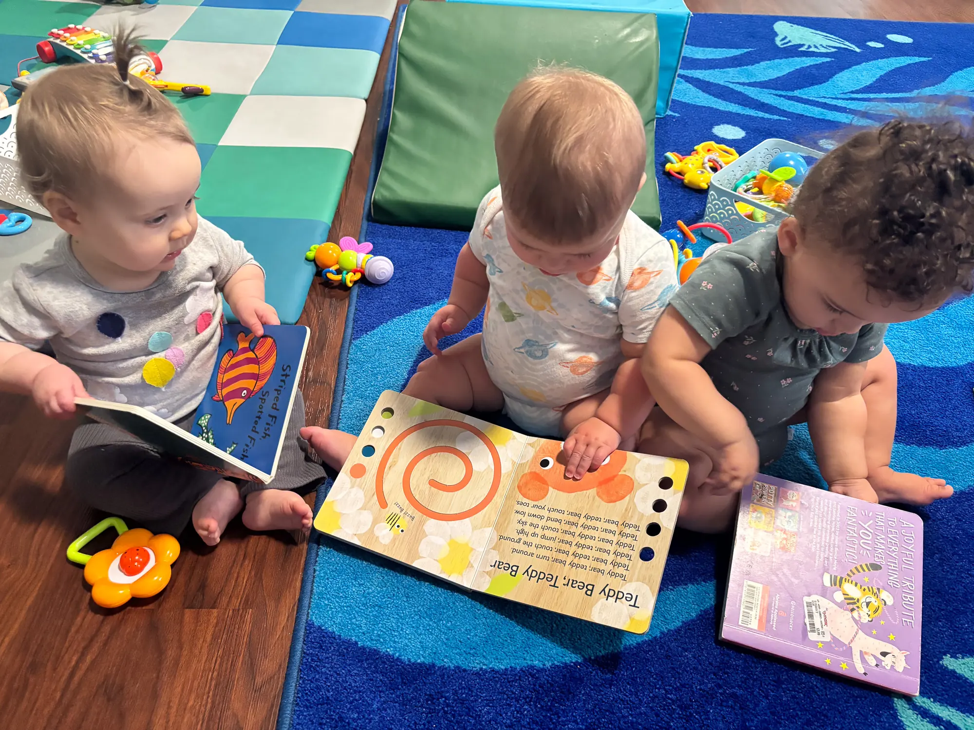Three babies sitting on a colorful play mat, exploring board books together