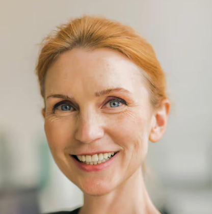 Close-up portrait of a smiling woman with light red hair and blue eyes against a soft-focused background.