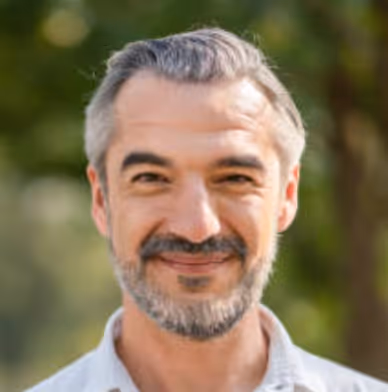Smiling middle-aged man with salt-and-pepper hair and beard standing outdoors with blurred green foliage background.