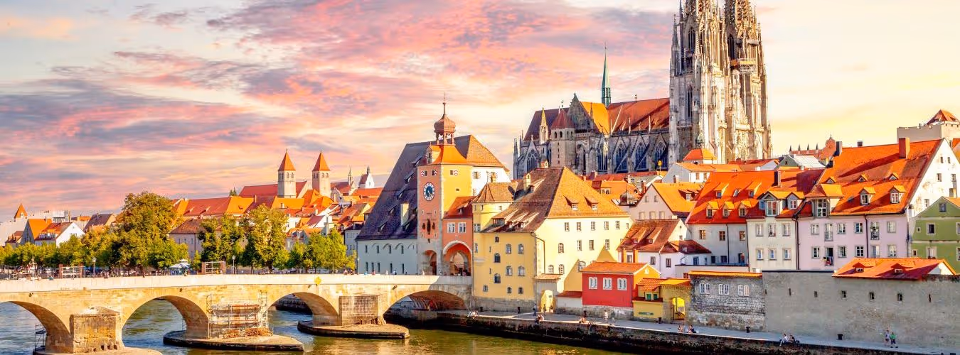 Historic European town with a stone bridge over a river, colorful buildings, and a large Gothic cathedral at sunset.