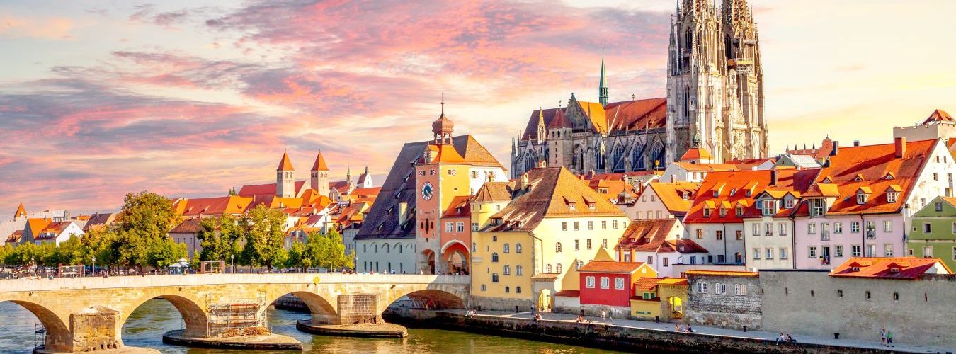 Historic European town with a stone bridge over a river, colorful buildings, and a large Gothic cathedral at sunset.