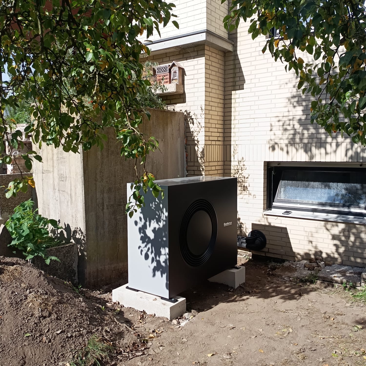 Schwarze Wärmepumpe neben Backsteinhaus mit Bäumen im Schatten
