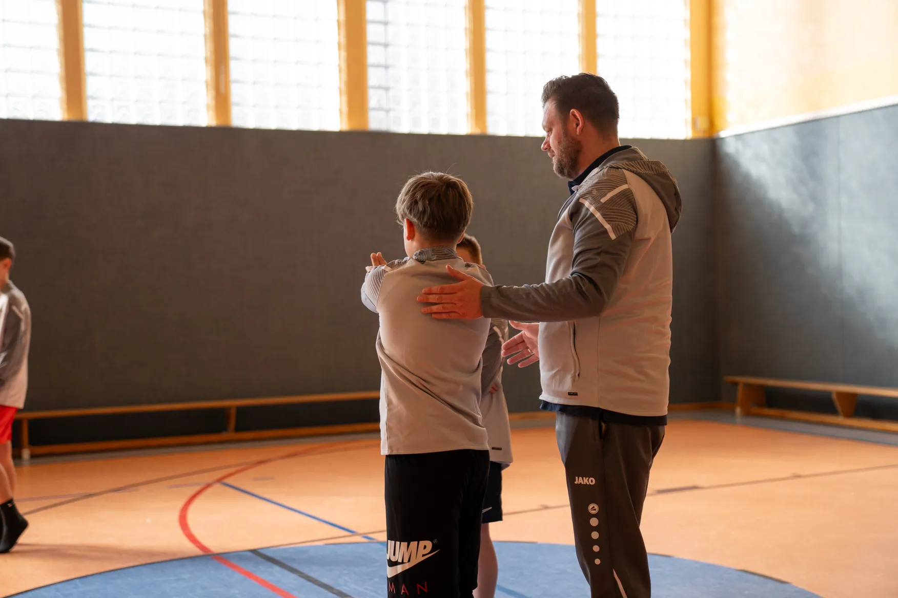 Coach instructing two boys in a gymnasium with wooden benches and large windows.