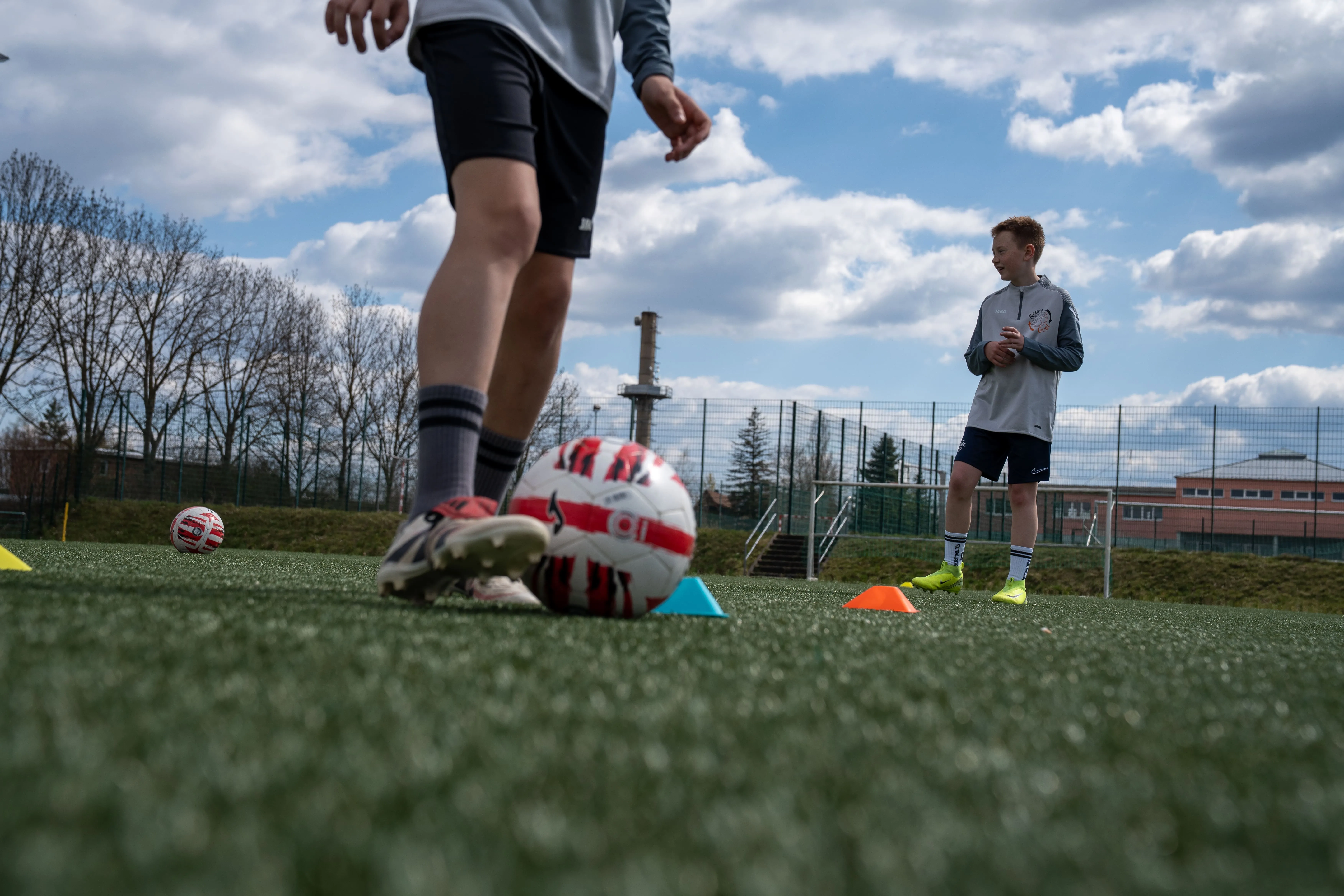 Two boys on a soccer field during practice with soccer balls, cones, and goalposts under a partly cloudy sky.