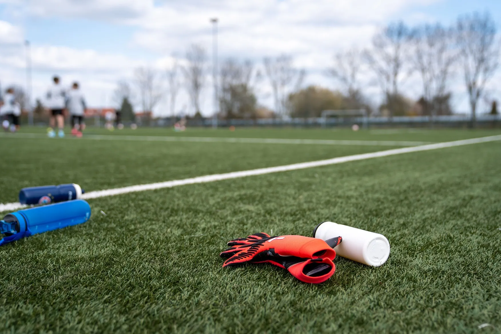 Red and black goalie gloves and a white water bottle lying on a green soccer field with players blurred in the background.