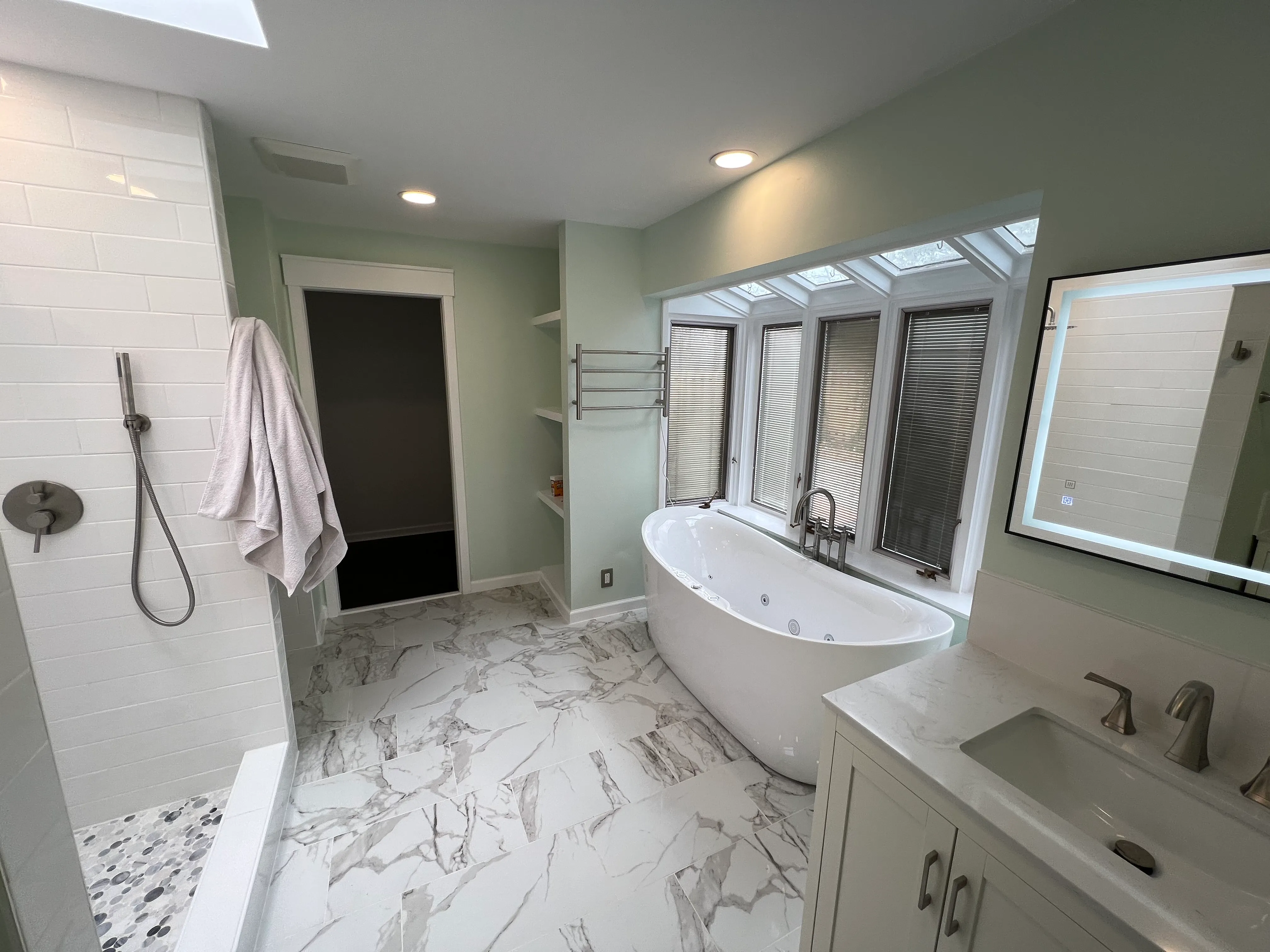 Modern bathroom with freestanding tub under angled windows, marble floor, walk-in shower, and vanity with lit mirror.