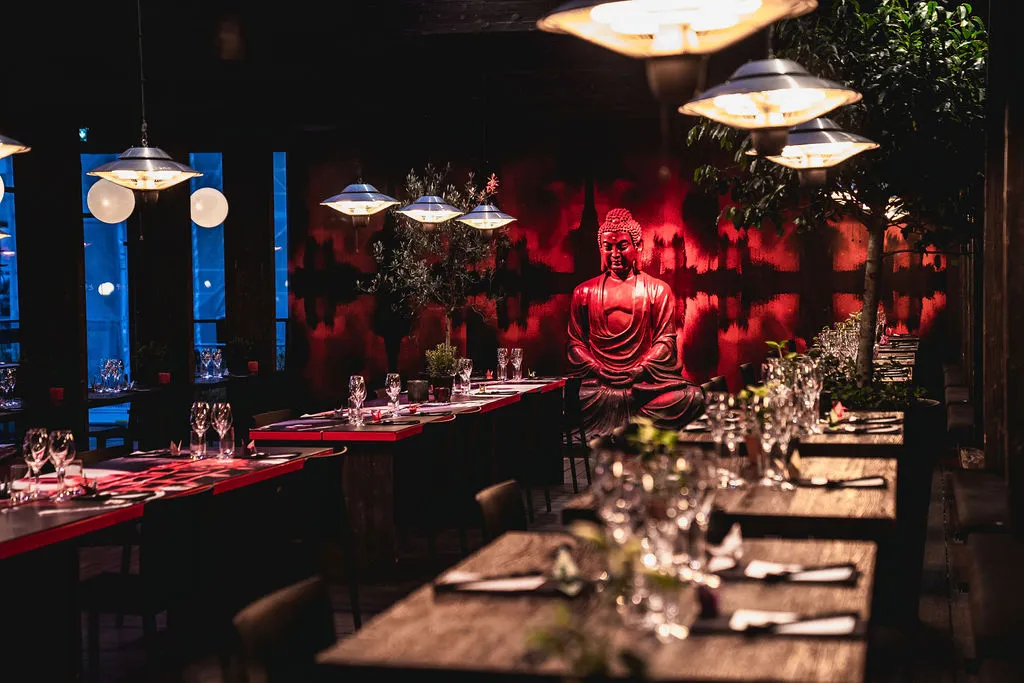 Dimly lit restaurant interior with wooden tables set with glassware, and a large red Buddha statue against a red and black patterned wall.