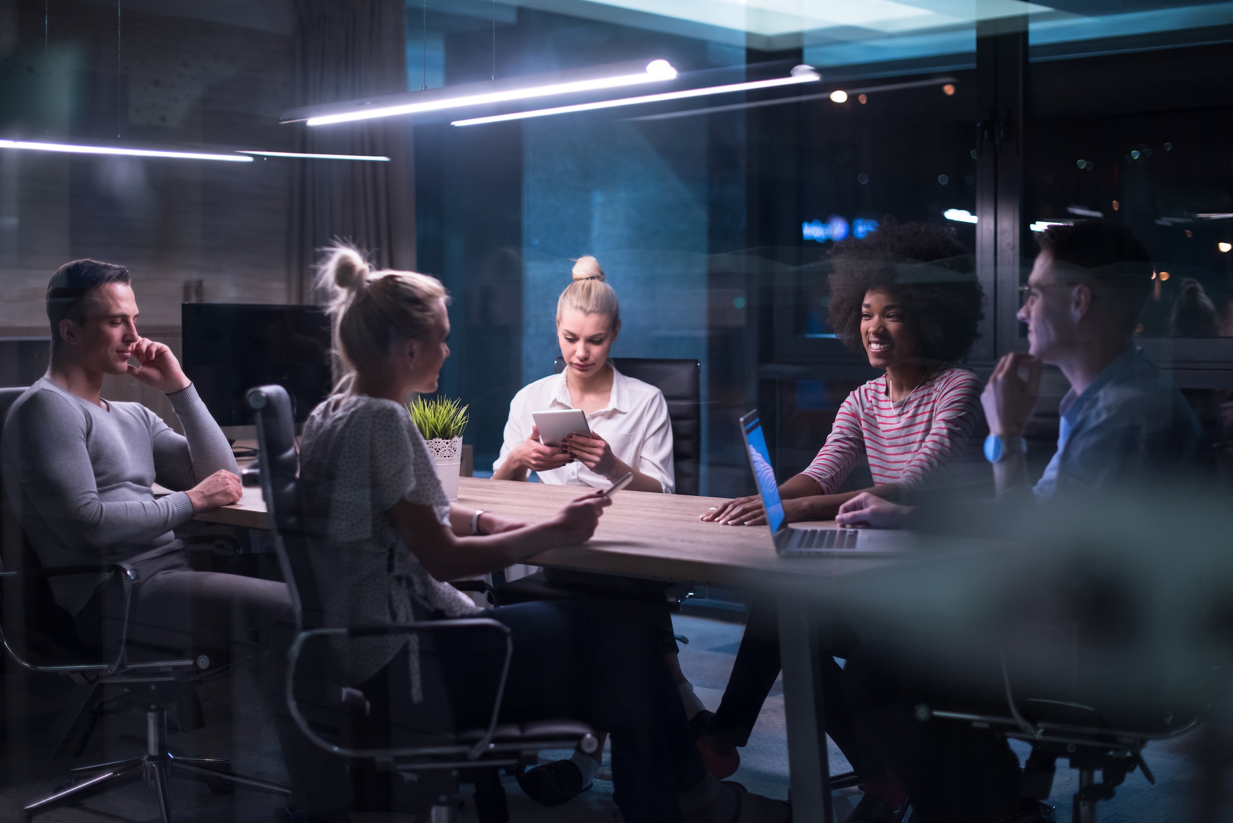 Five diverse colleagues having a night meeting in a modern office with laptops and tablets.