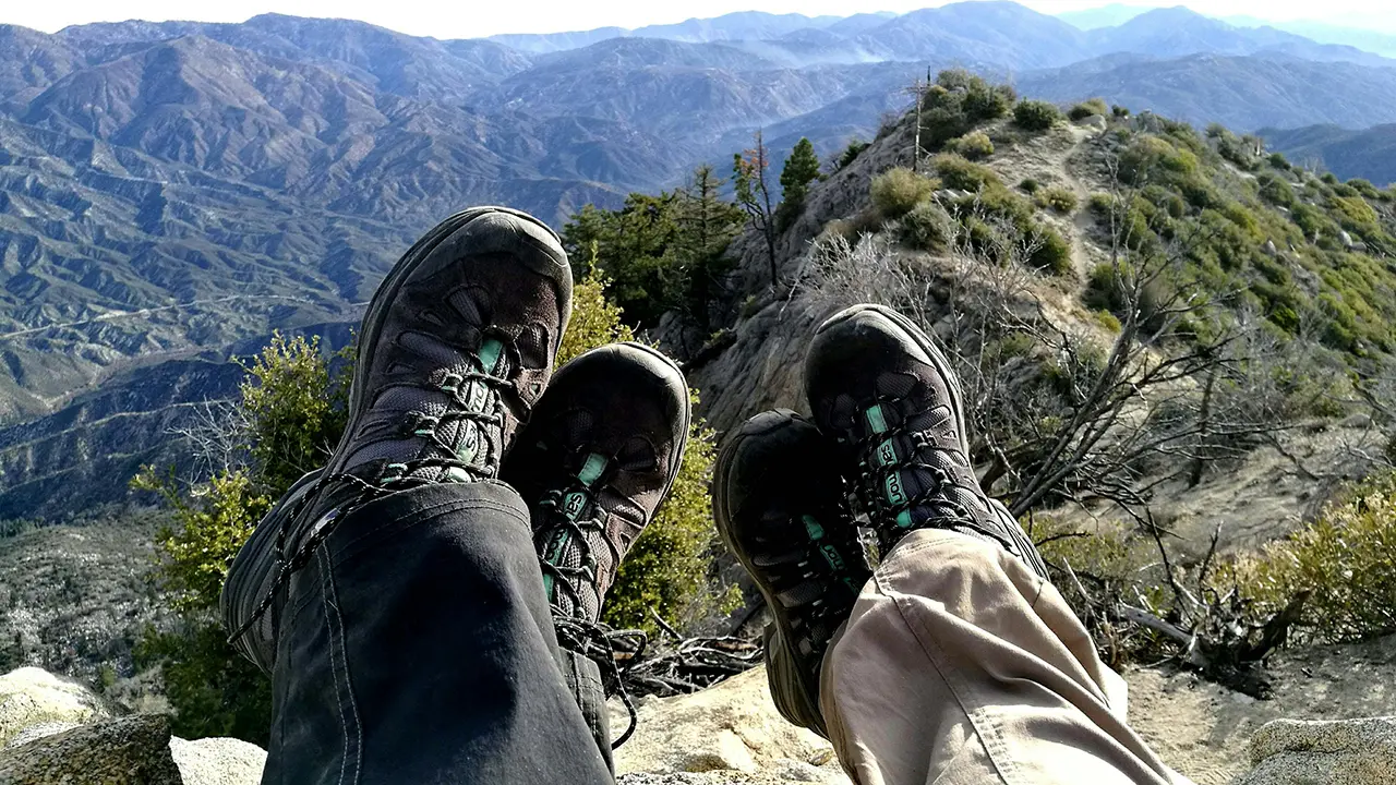 Two hikers resting with their boots stretched out toward a panoramic mountain landscape and rugged outdoor terrain.