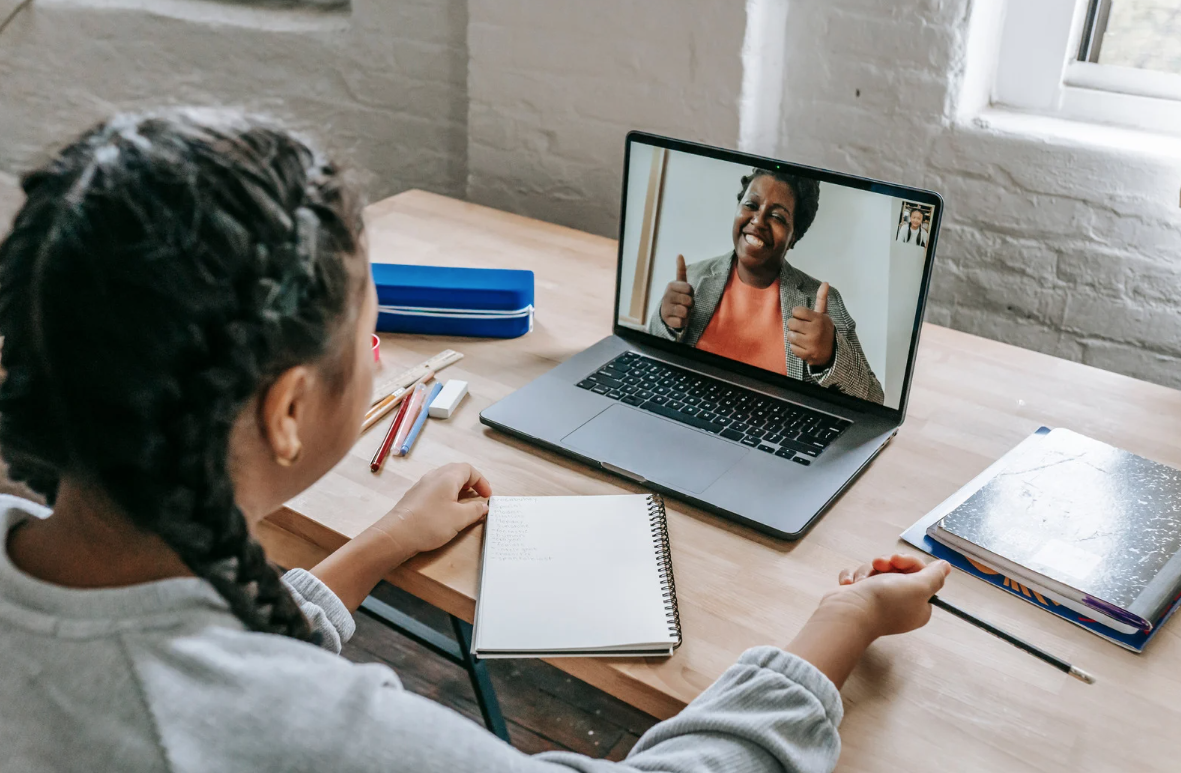 Child sits at desk with notebook, video chatting on laptop where woman gives thumbs-up.