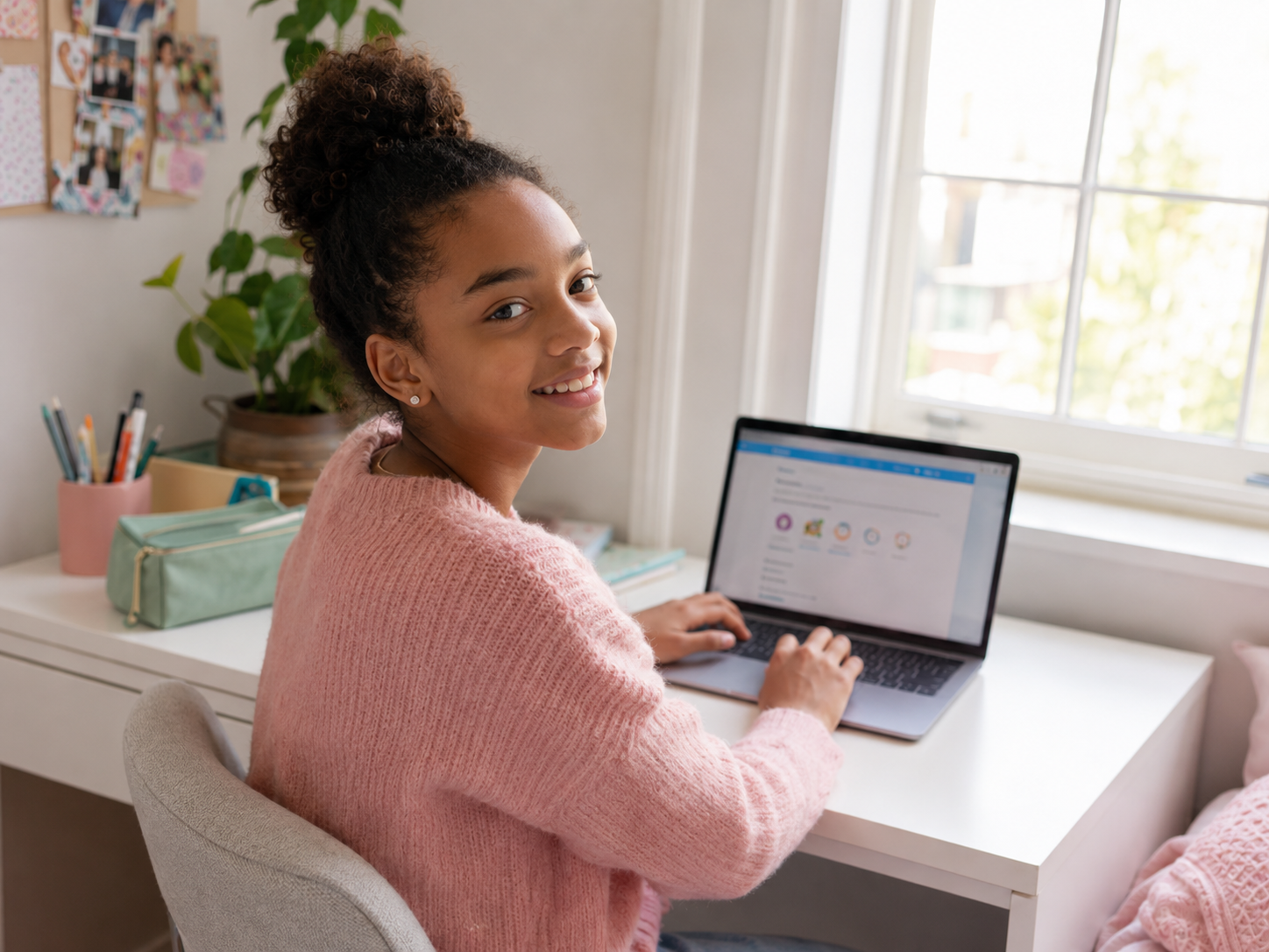 Smiling girl in a pink sweater using a laptop at a white desk near a window during the day.