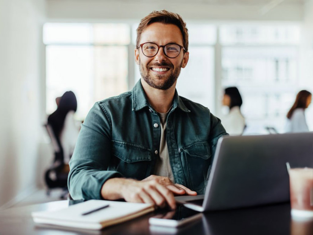 Smiling man with glasses working on a laptop at a desk with a notebook and phone in a bright office.