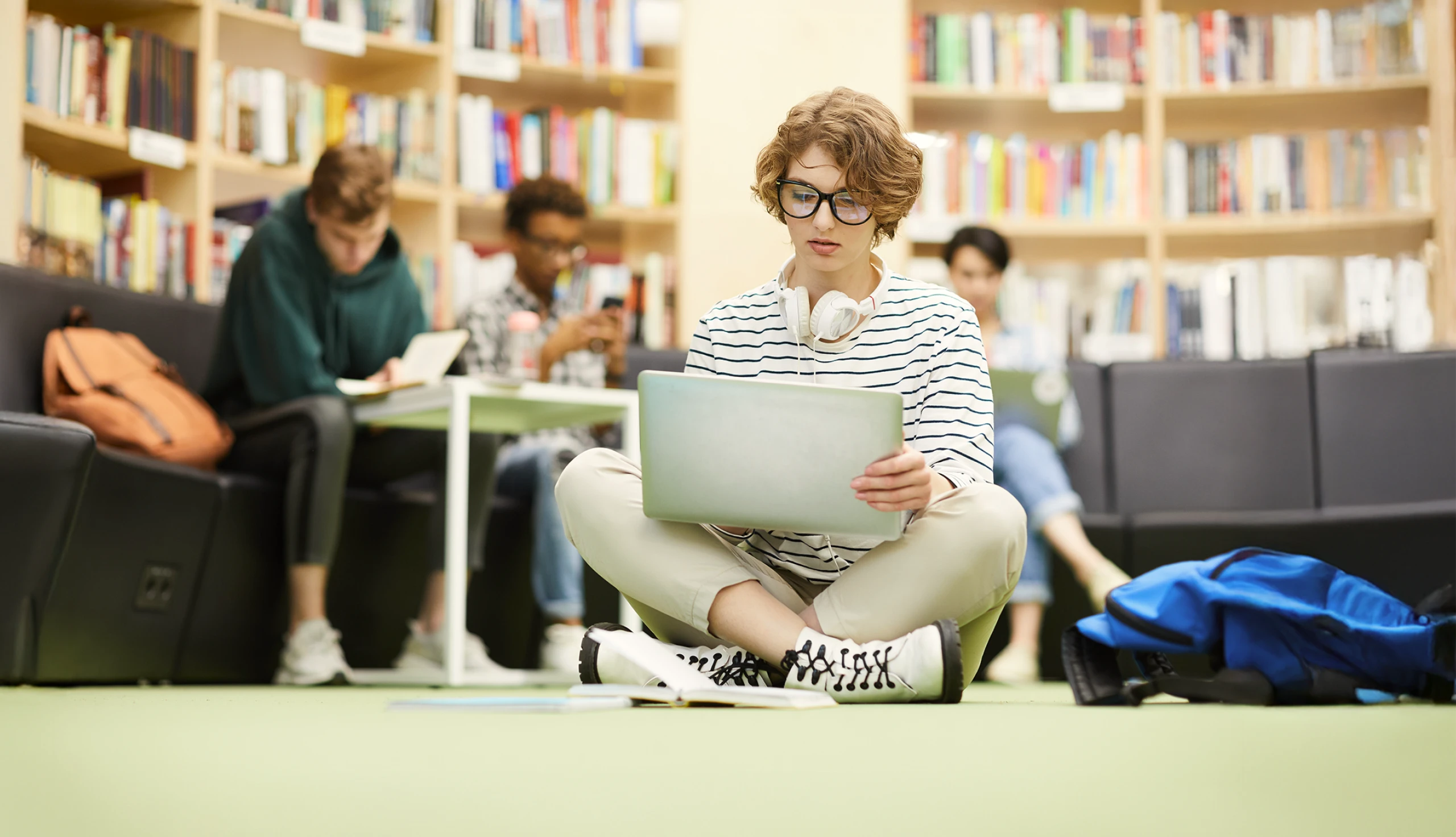 A student sitting on the floor in a library working on their laptop with other students in the background.