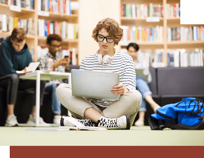 A student sitting on the floor in a library working on their laptop with other students in the background.