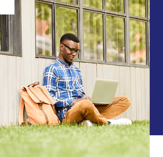 A student working on a laptop, sitting on grass and leaning against a building with his backpack beside him.
