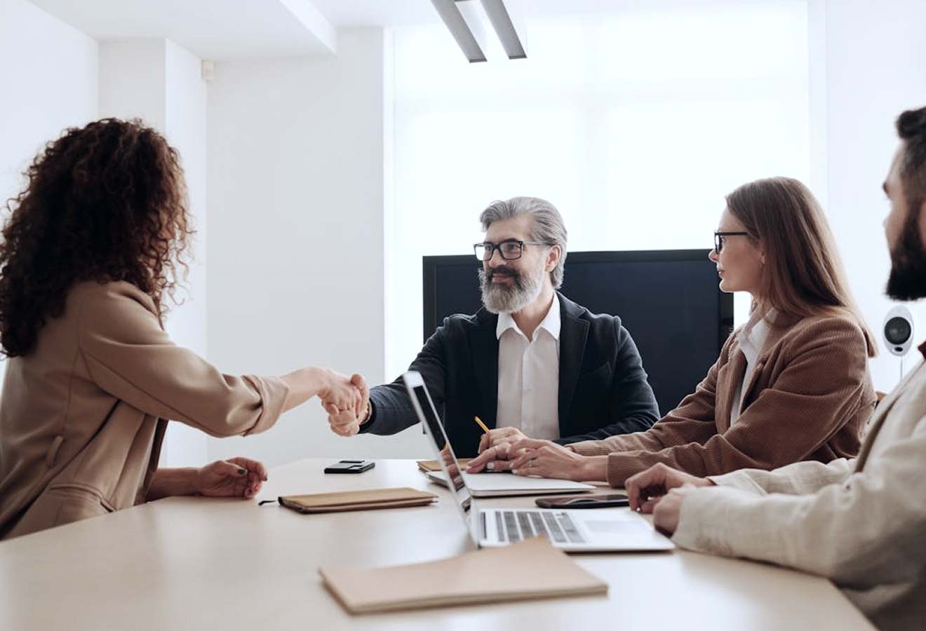 People in a business meeting shaking hands at a conference table with laptops and documents.