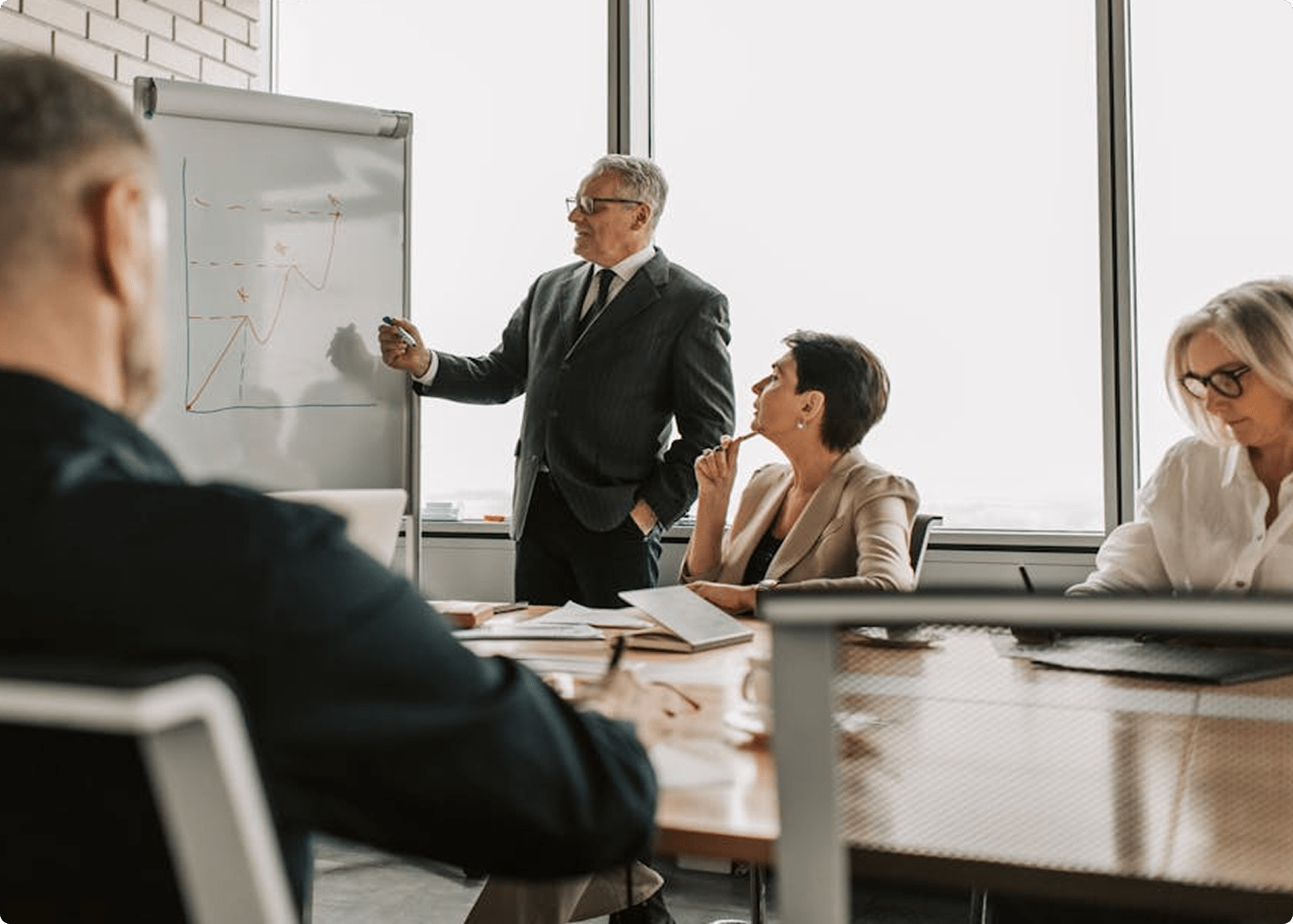 Businessman giving a presentation using a whiteboard with graphs while colleagues listen and take notes in a modern office.