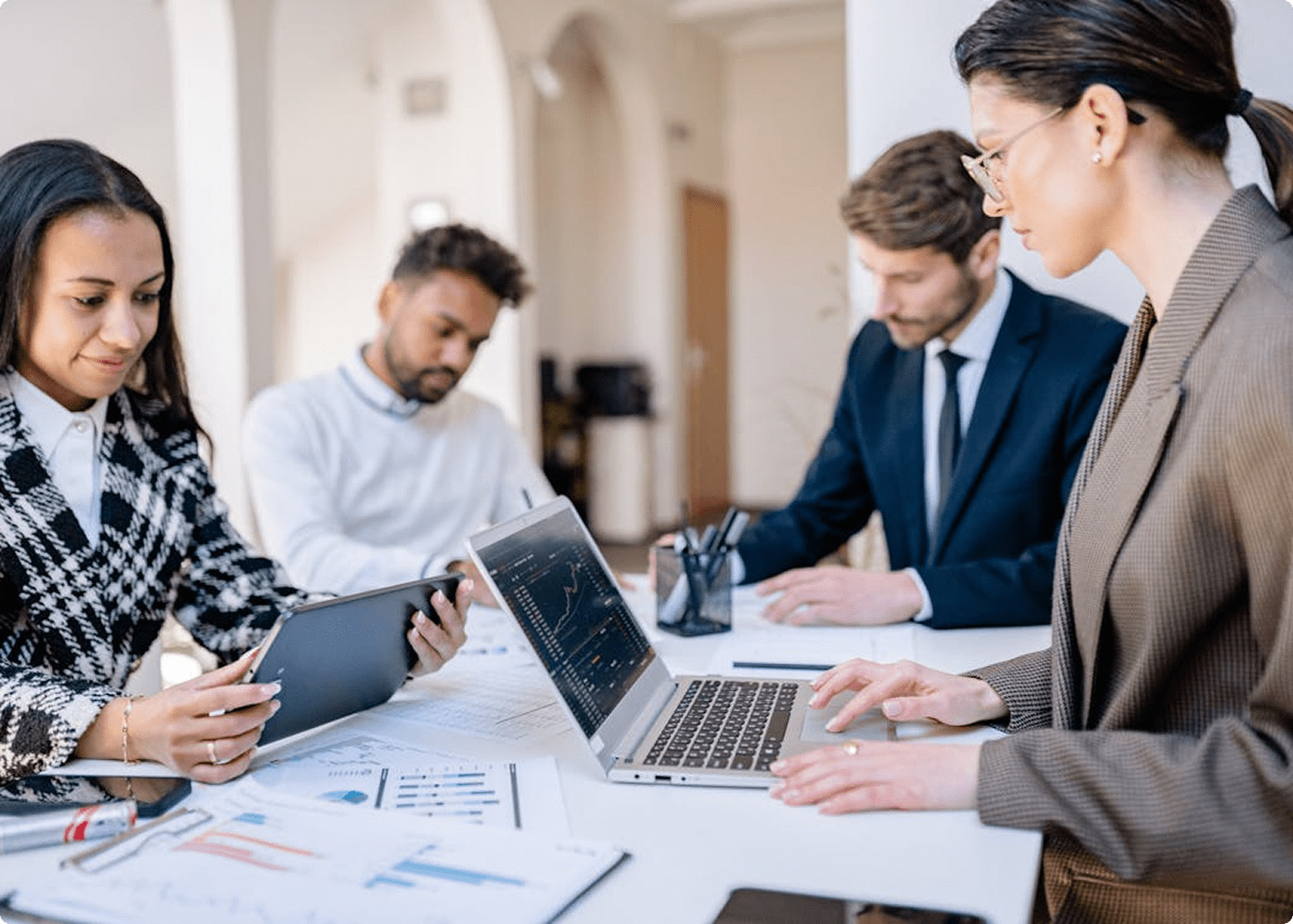 Four diverse professionals working together at a table with a laptop, tablet, and printed charts in a bright office.