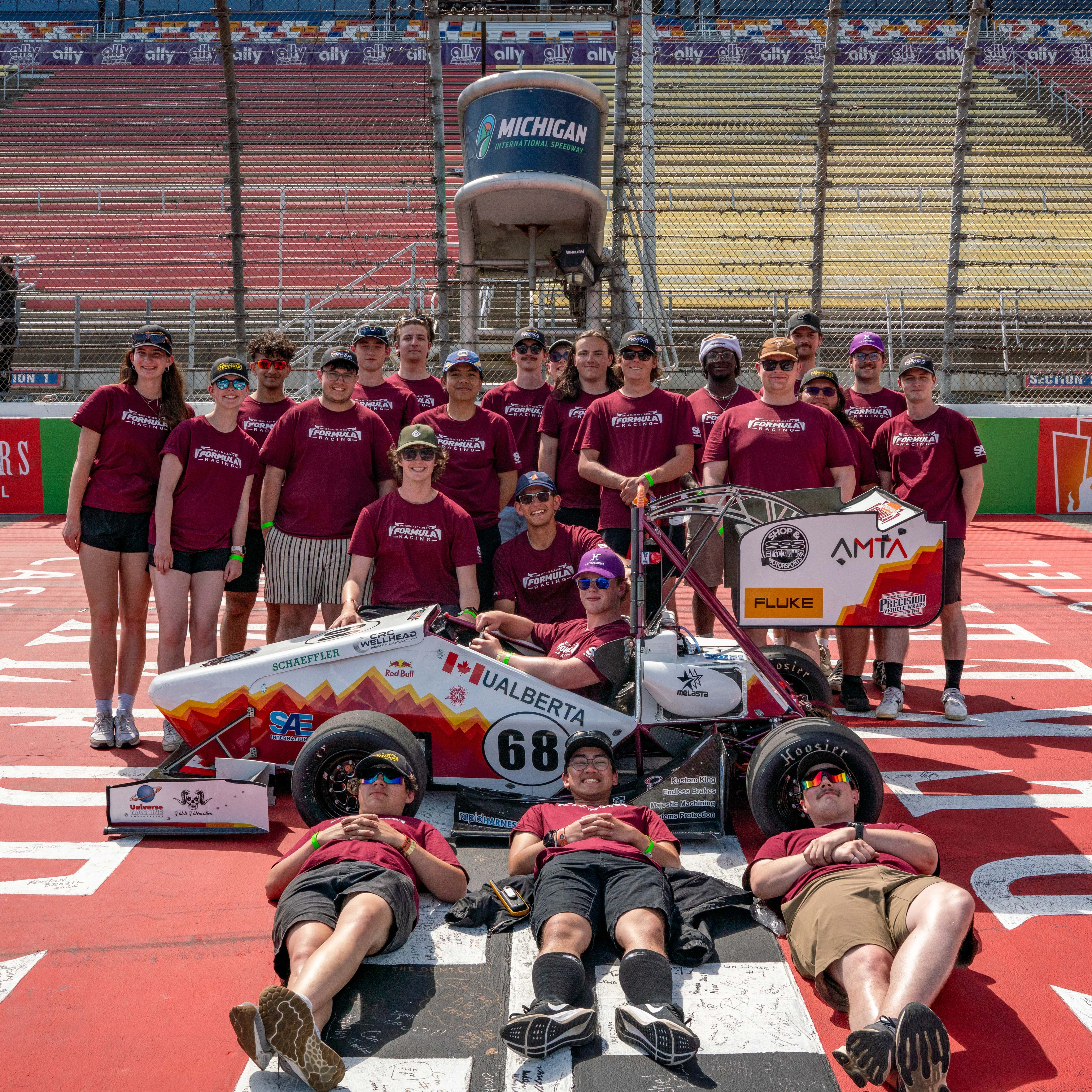 Team in matching shirts poses with race car numbered 68 on a racetrack near Michigan Speedway sign.