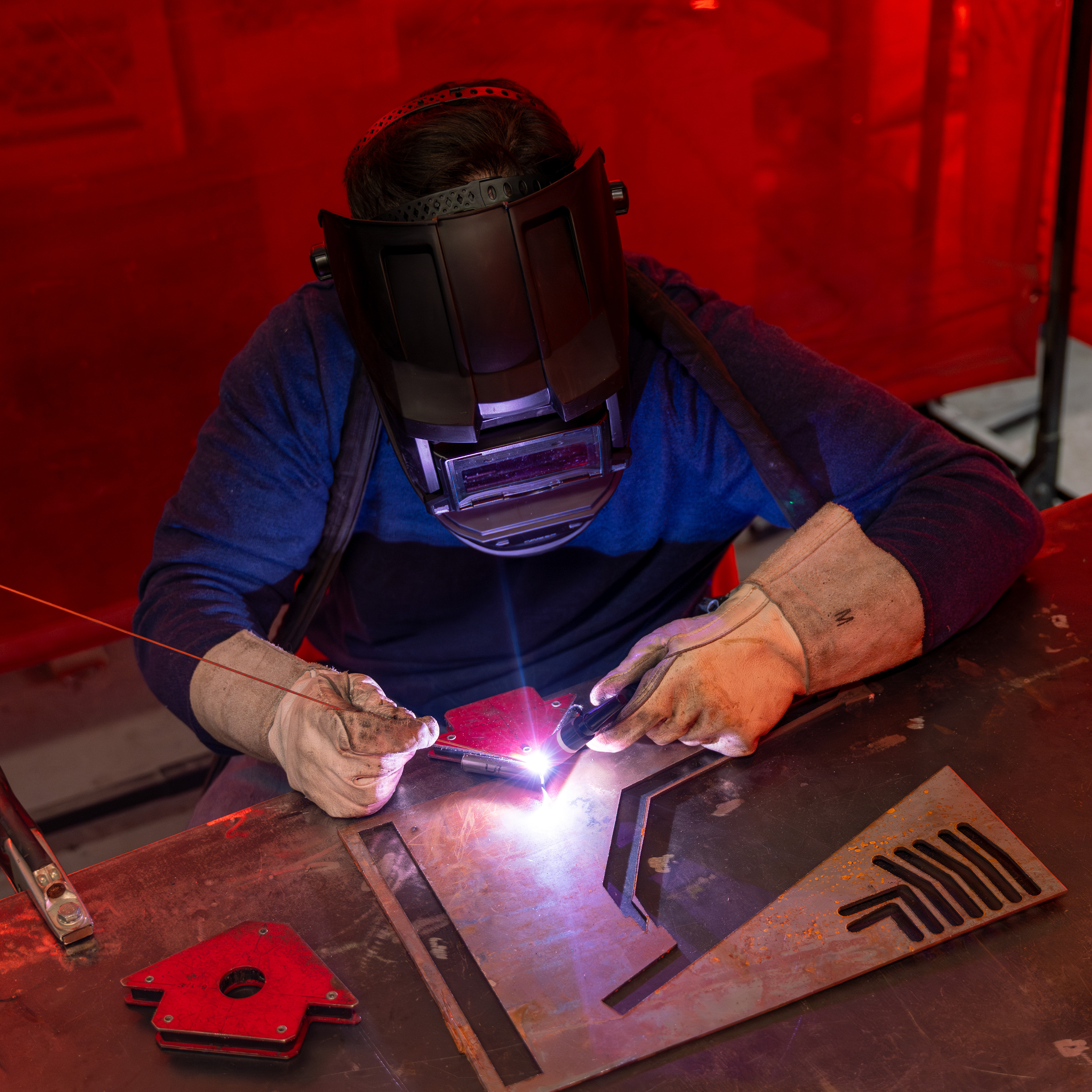 Worker wearing a welding helmet and gloves while welding metal pieces on a table with red background.