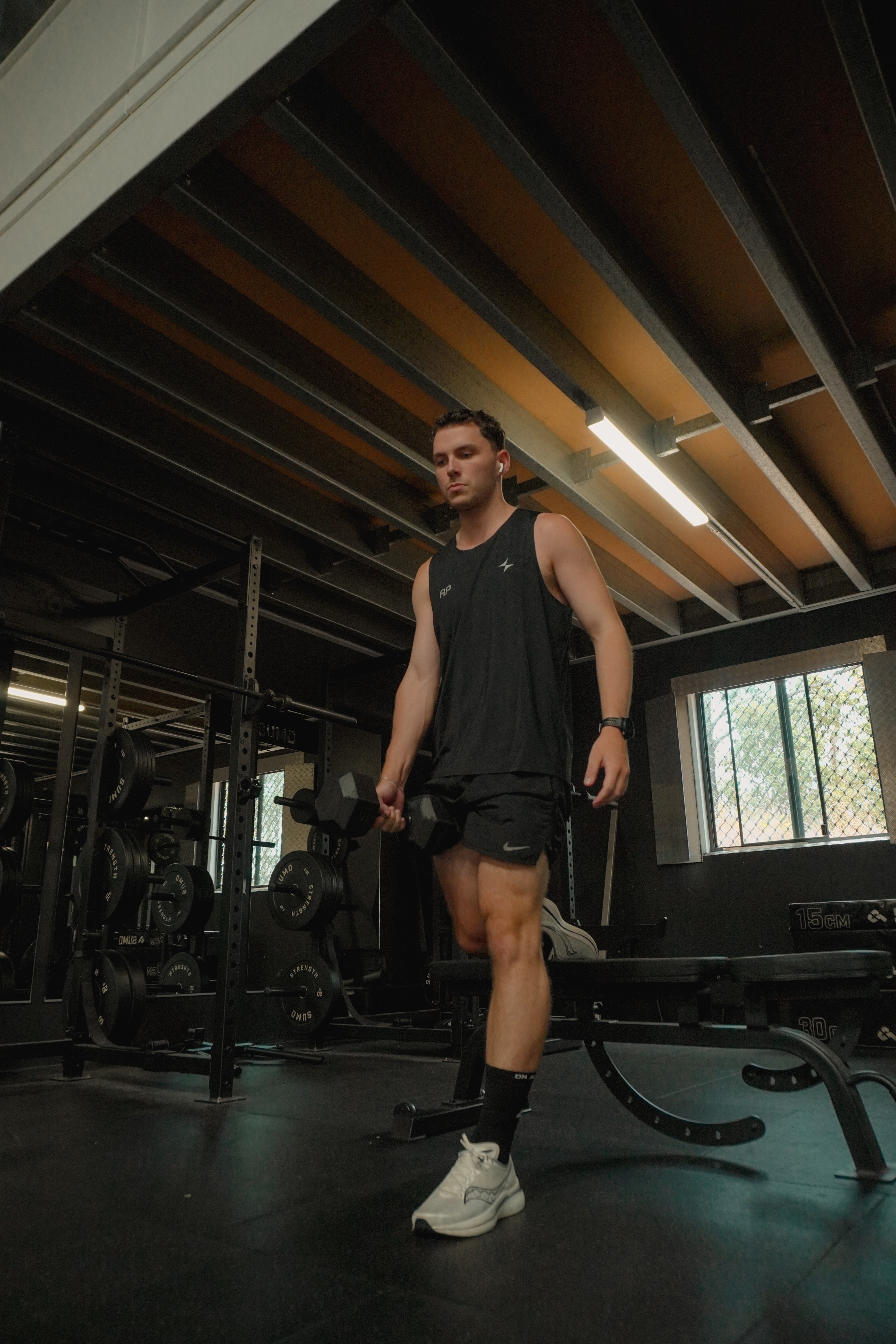 Young man in black athletic wear walking in a gym holding a dumbbell in each hand.