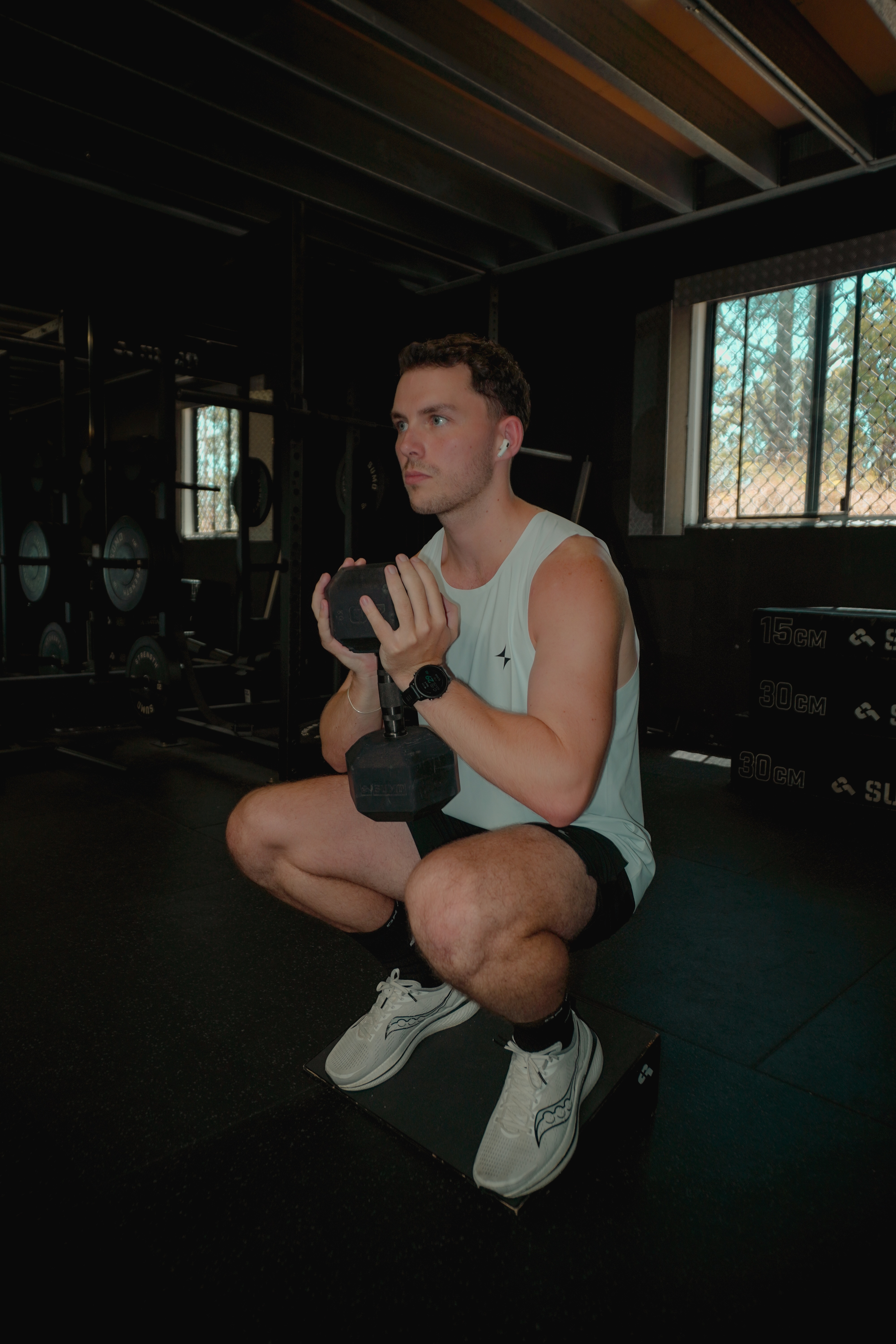 Man in a white tank top and black shorts performing a dumbbell squat on a box in a gym.