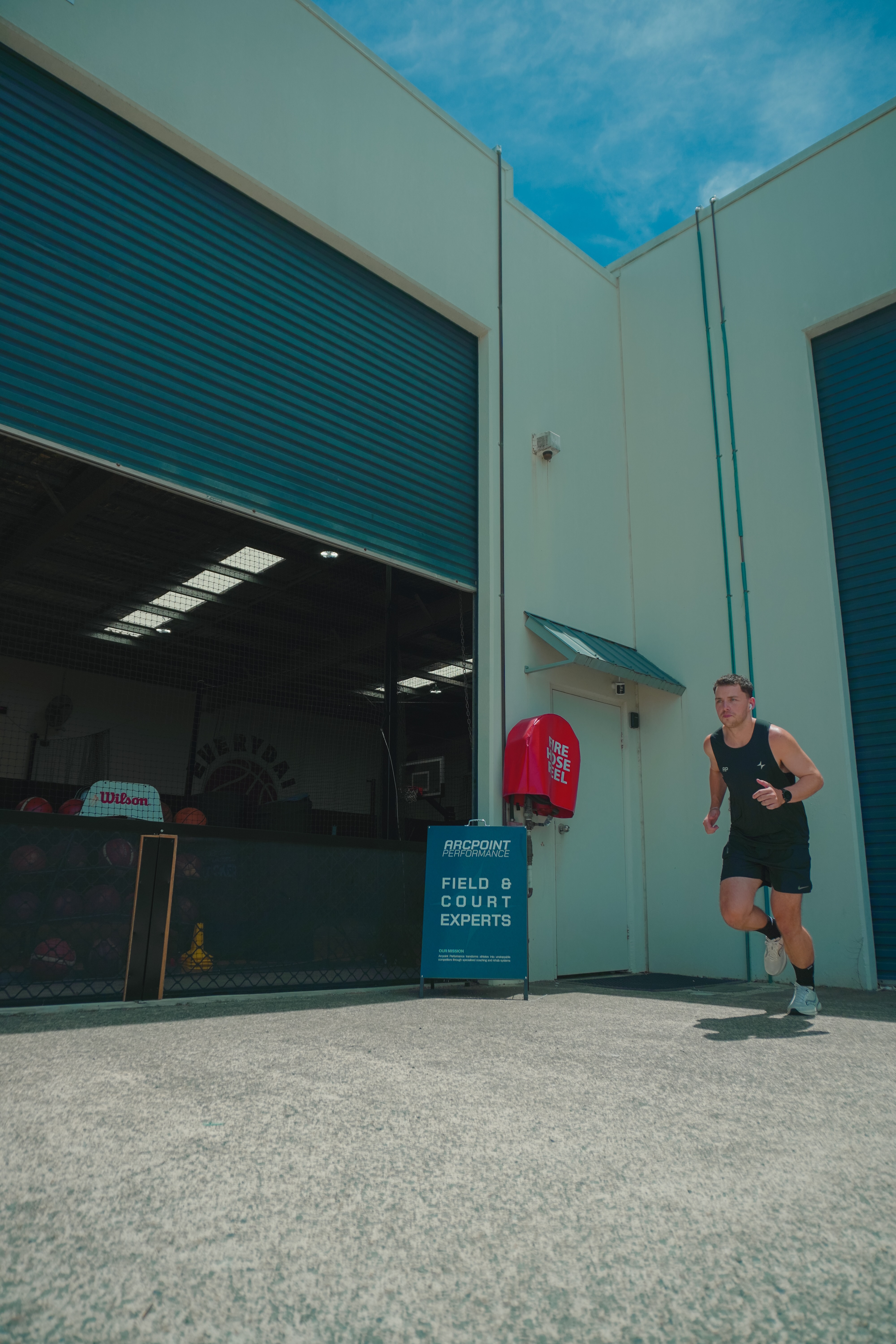 Man in black athletic wear running outside a sports facility with a partially open roller door and a sign reading 'Field & Court Experts'.