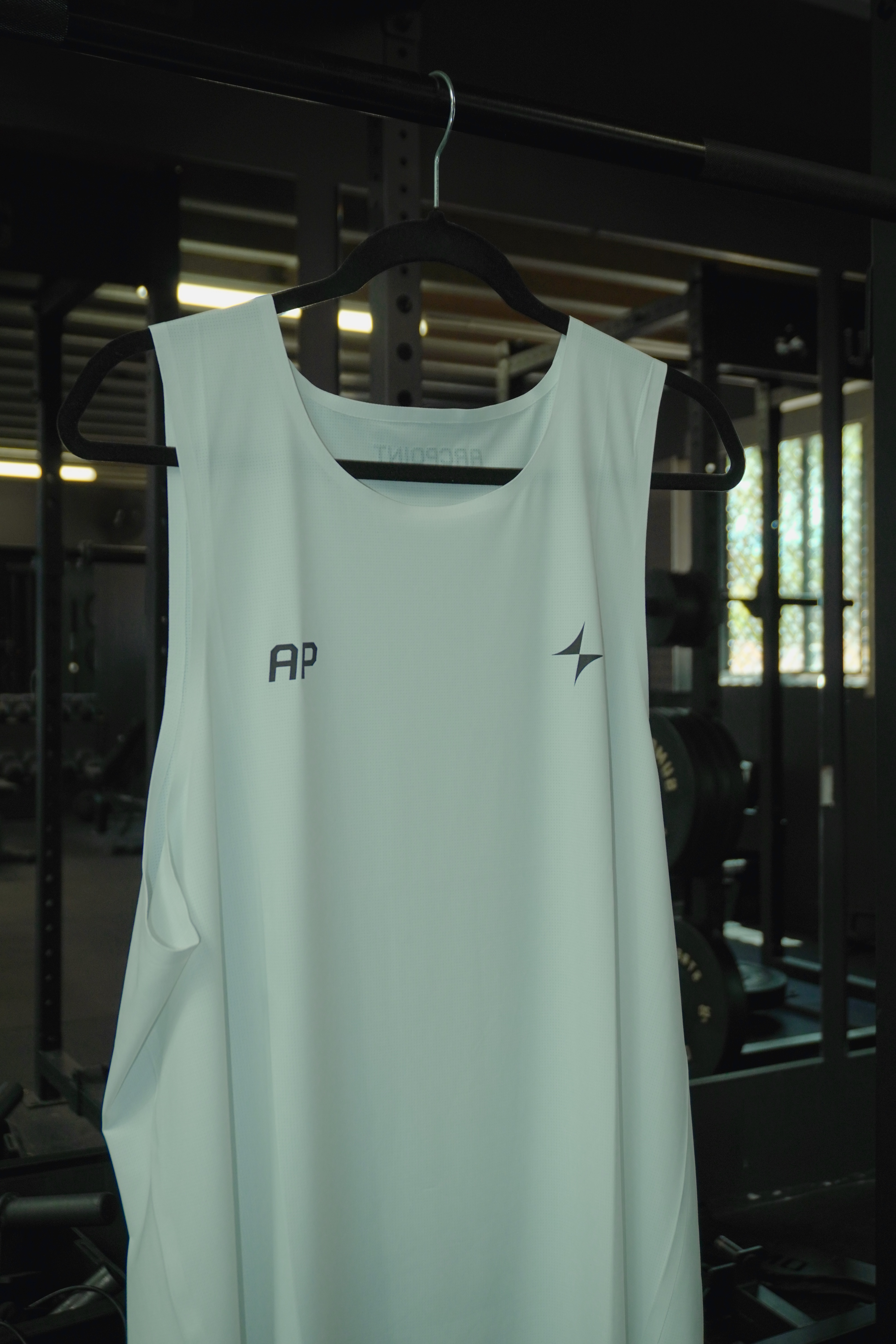 White sleeveless athletic shirt with AP logo and Arc symbol hanging on a black hanger in a gym.