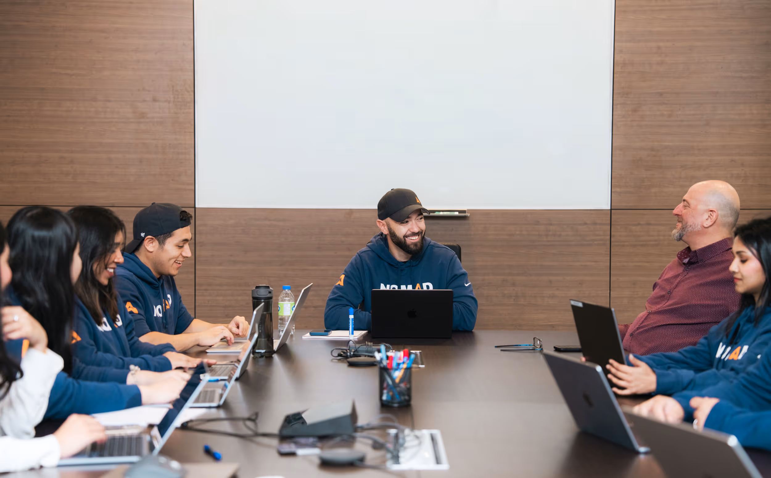 A diverse group of people in hoodies and casual wear sitting around a conference table with laptops, engaged in a meeting and smiling.
