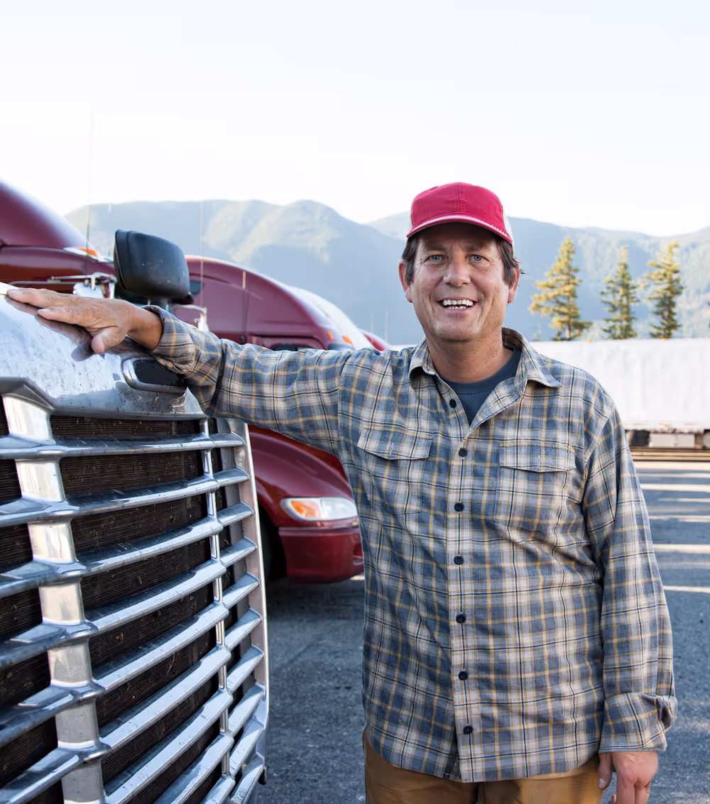Smiling man in a red cap and plaid shirt standing next to a large truck with mountains in the background.