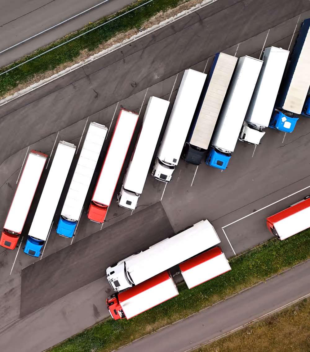 Aerial view of a parking lot with multiple large semi-trucks and trailers parked in angled spaces, mostly white trailers with red and blue cabs.