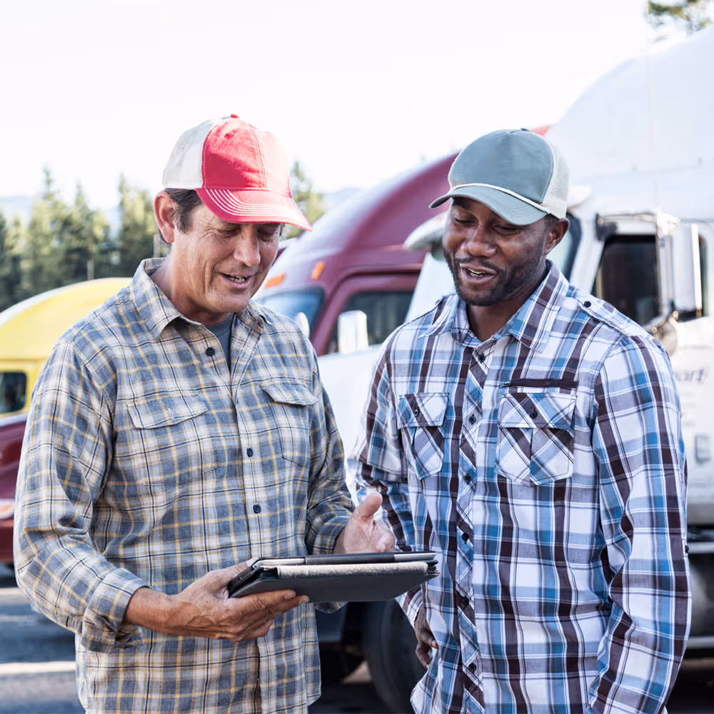 Two men in plaid shirts and caps looking at a tablet, standing in front of parked trucks.