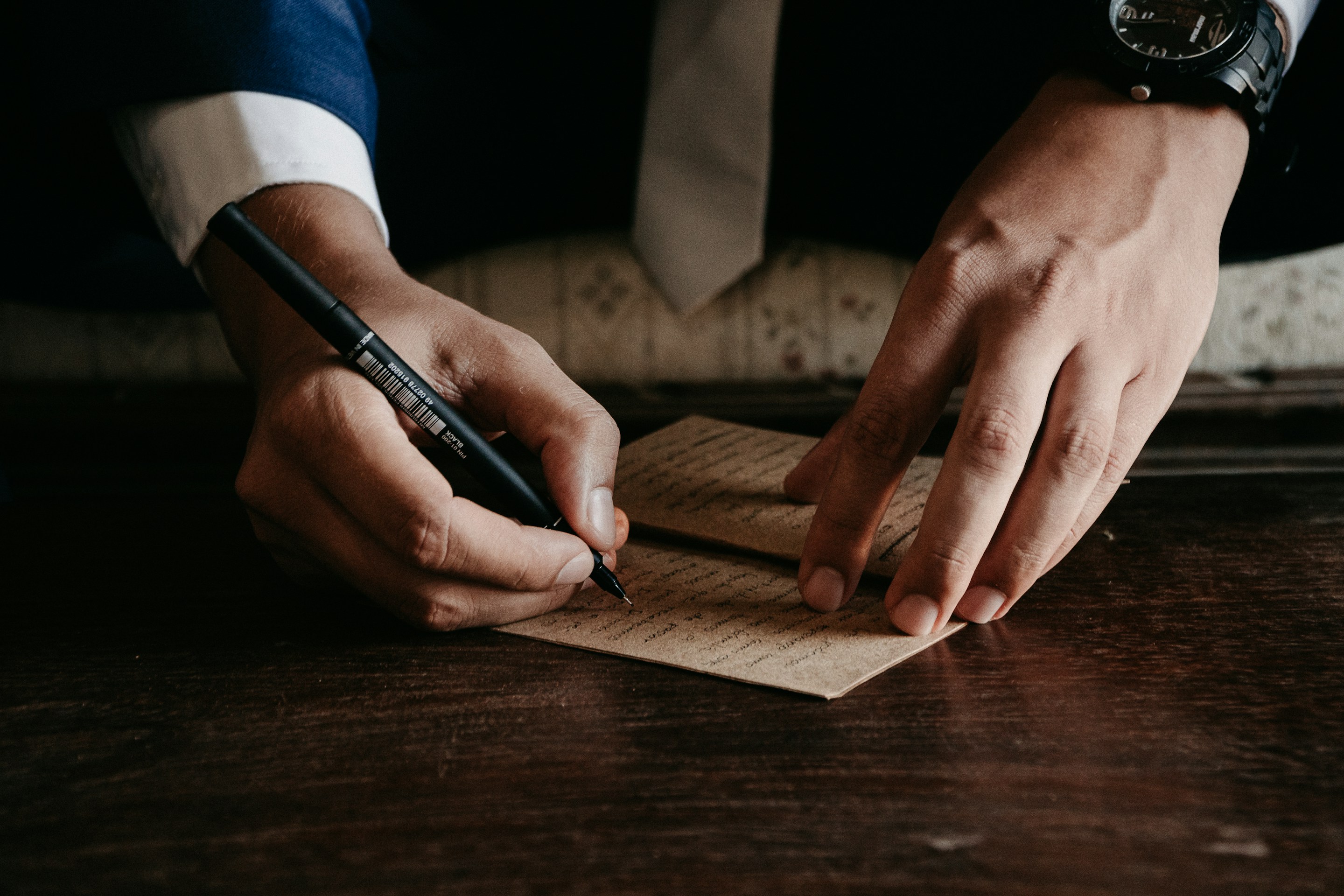 Person wearing a suit and tie writing on brown paper with a black pen on a wooden table.