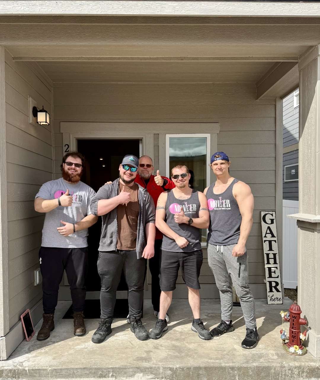 Five men standing on a porch in front of a house, four are giving thumbs up, two wear matching gray tank tops with a pink heart logo, and a vertical sign to the right reads 'GATHER here.'