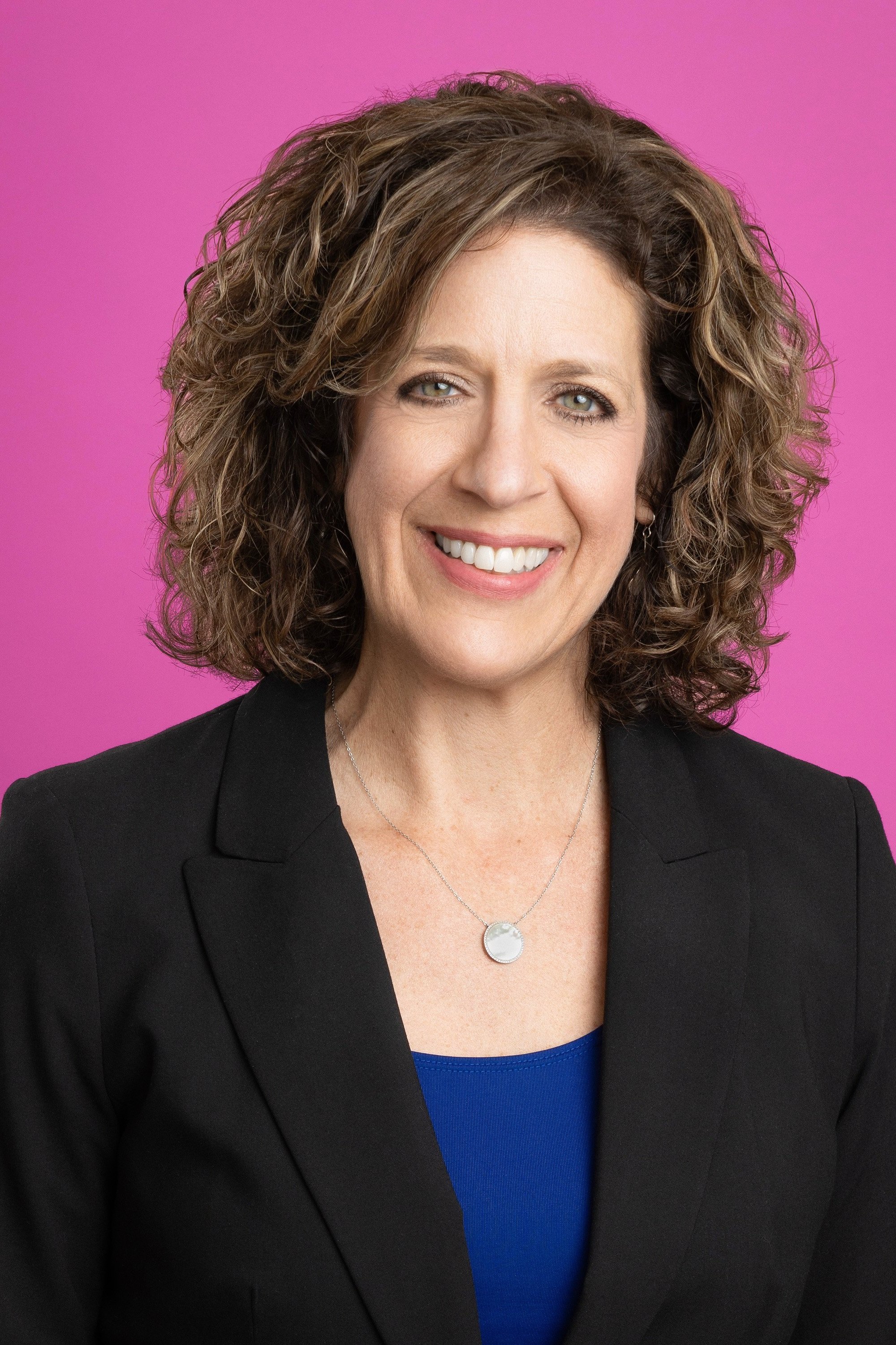 Smiling woman with curly brown hair wearing a black blazer over a blue top and a silver pendant necklace, against a pink background.