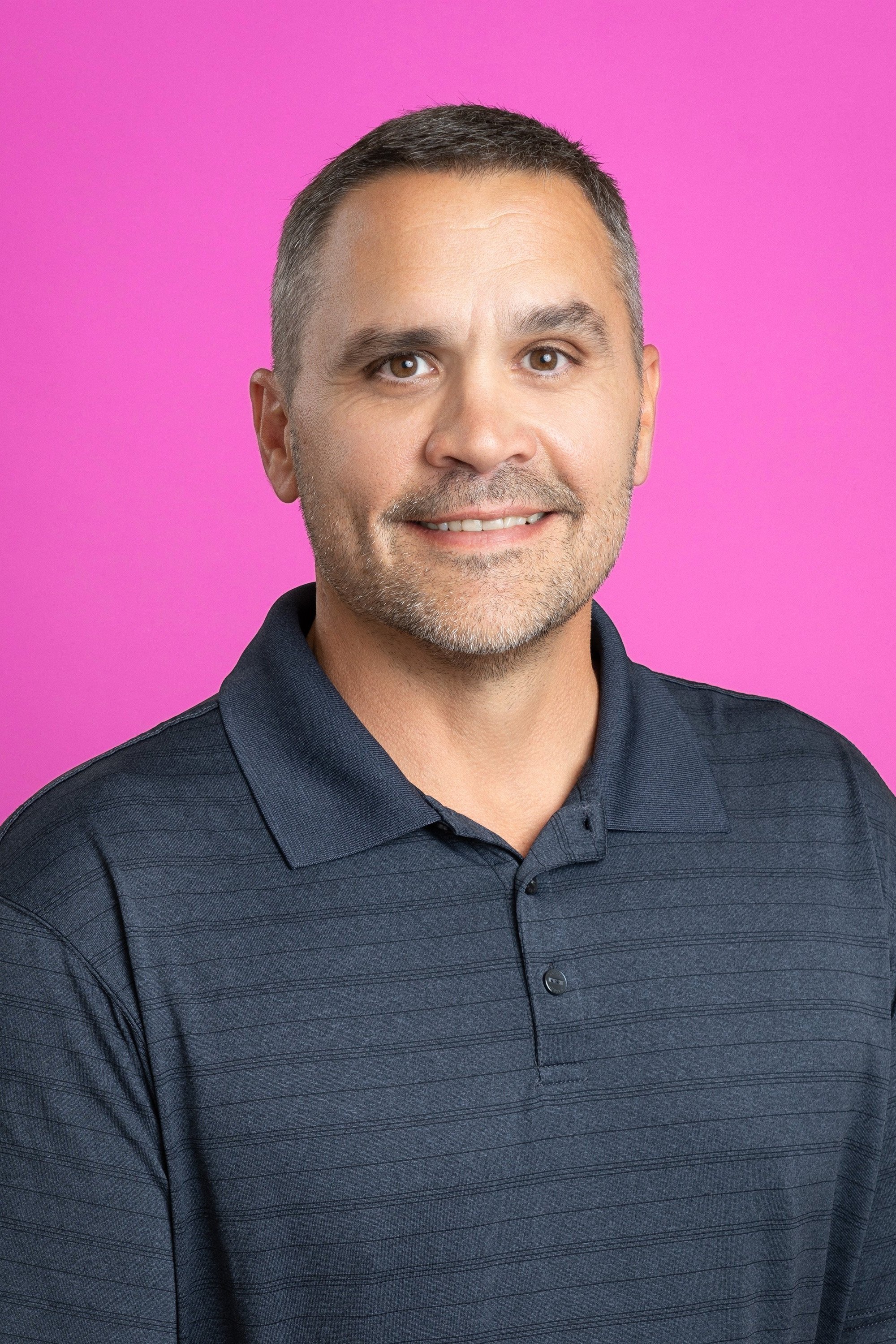 Smiling man with short dark hair and stubble wearing a dark blue polo shirt against a bright pink background.