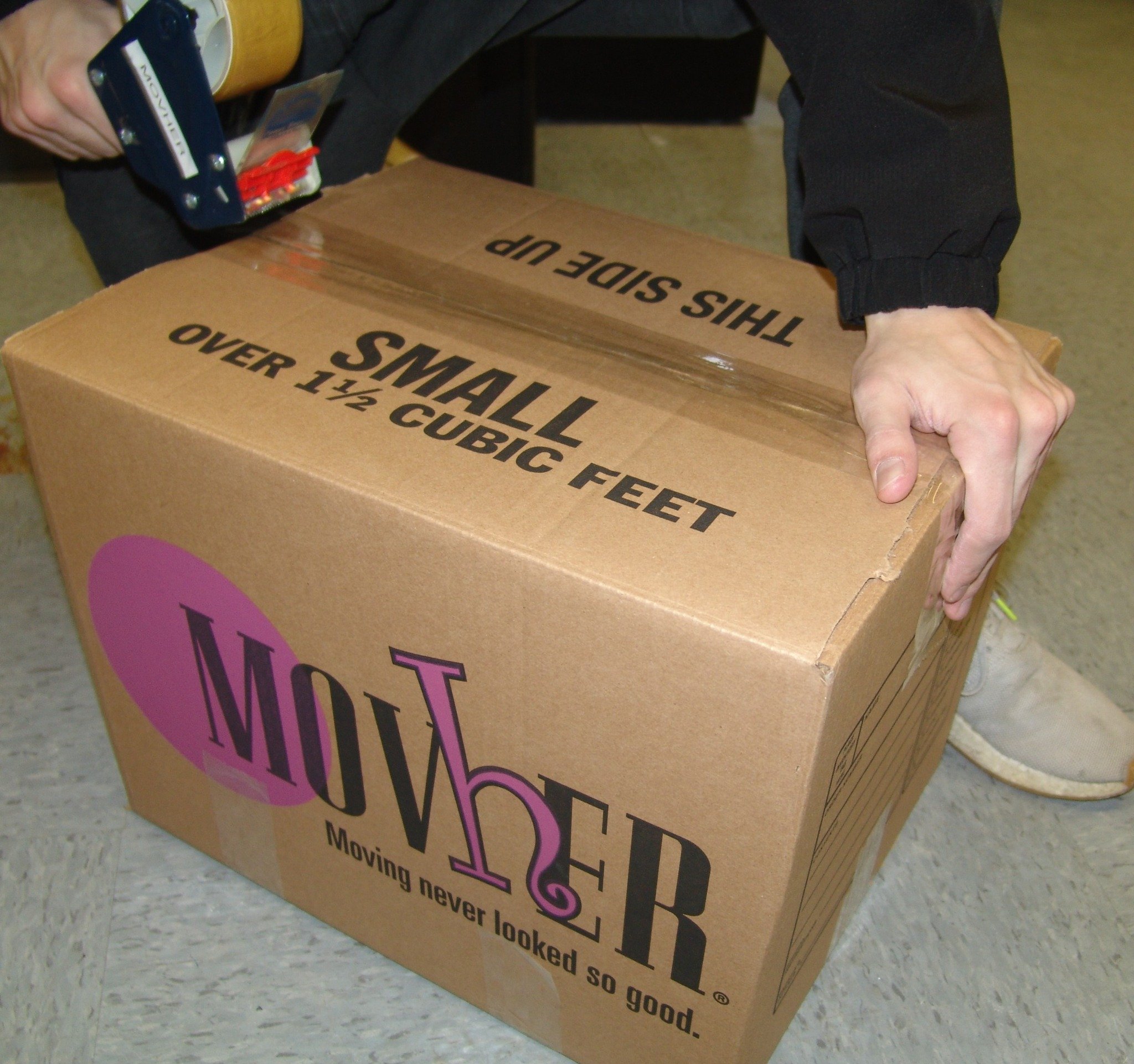 Person sealing a cardboard box labeled 'SMALL OVER 1 1/2 CUBIC FEET' with tape.