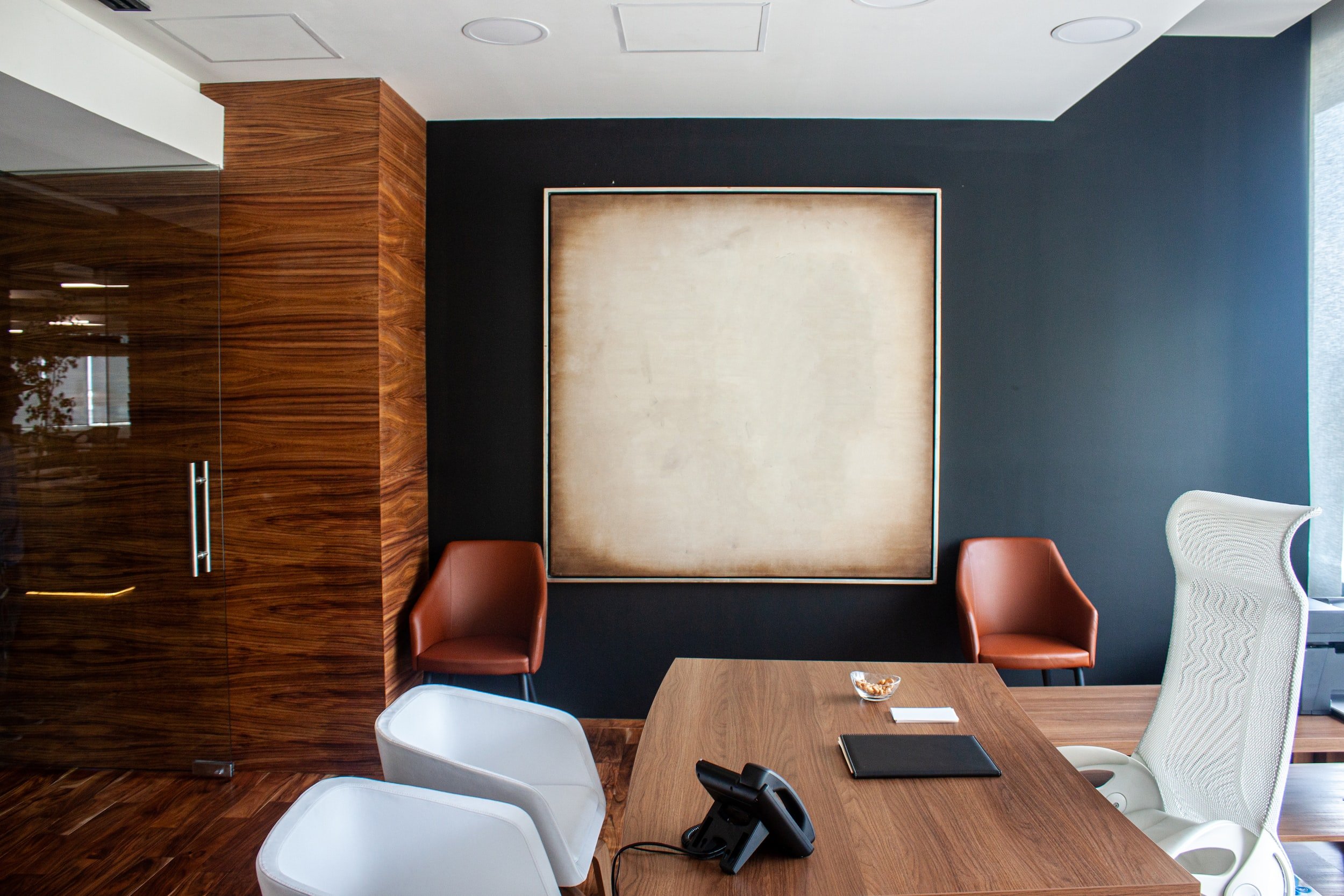 Modern office interior with a wooden desk, white ergonomic chair, two white guest chairs, two brown leather visitor chairs, and a large framed blank beige artwork on a dark wall.