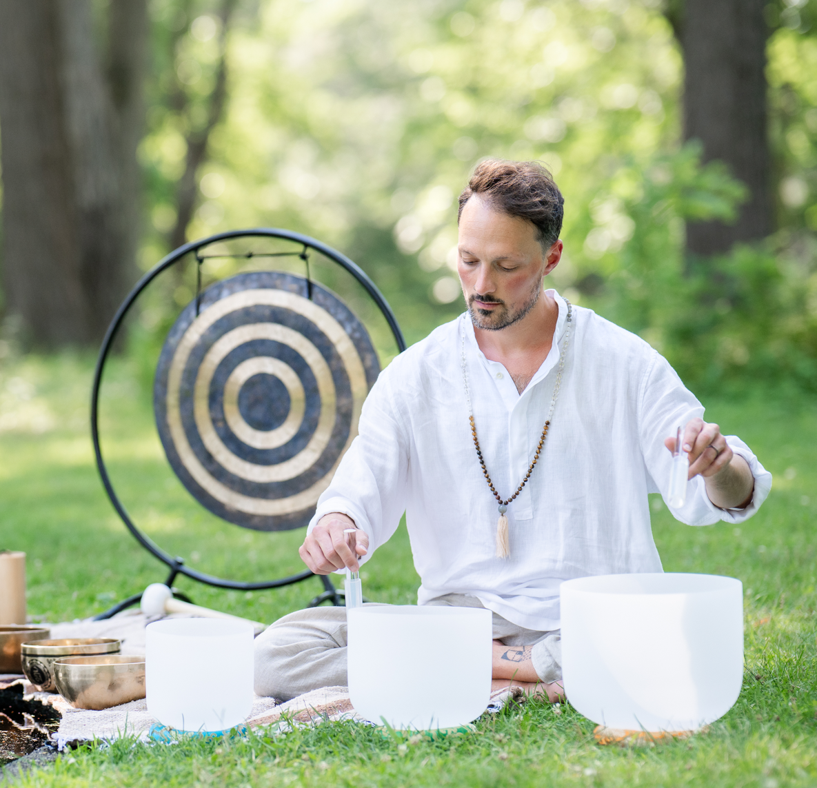 Matias in white shirt playing crystal singing bowls outdoors with a gong in the background.