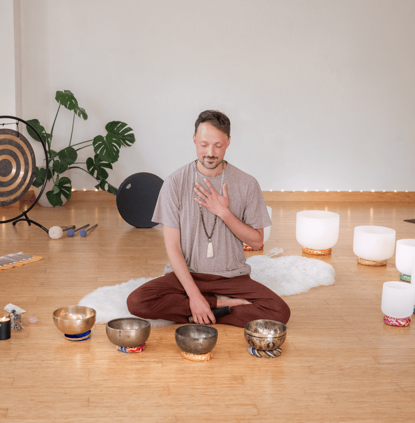 Matias sitting cross-legged on wooden floor surrounded by singing bowls and crystal bowls, with eyes closed and hand on chest in meditation.