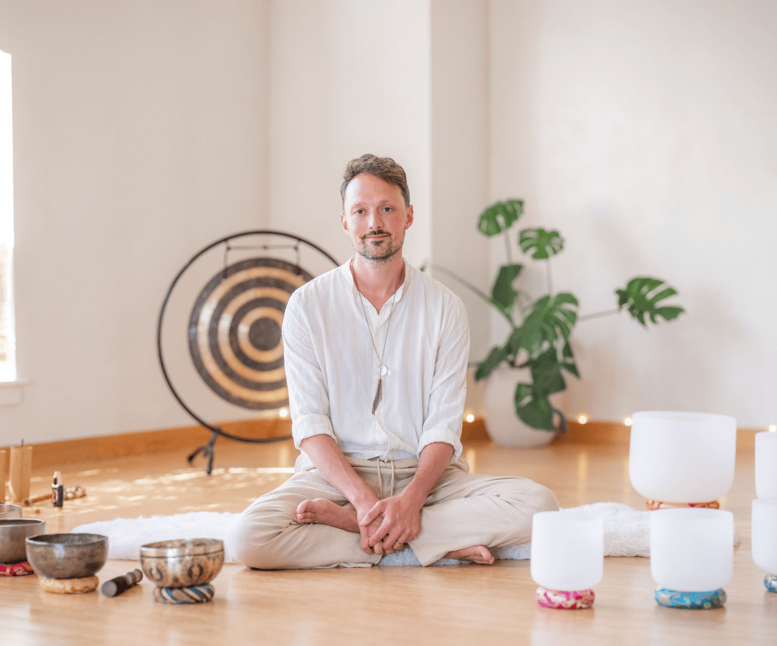 Matias in white shirt sitting cross-legged on floor surrounded by singing bowls and a gong in a bright room.