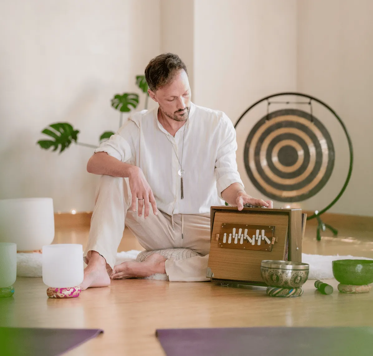 Matias seated cross-legged on a yoga mat holding a singing bowl and mallet in a sunlit room.