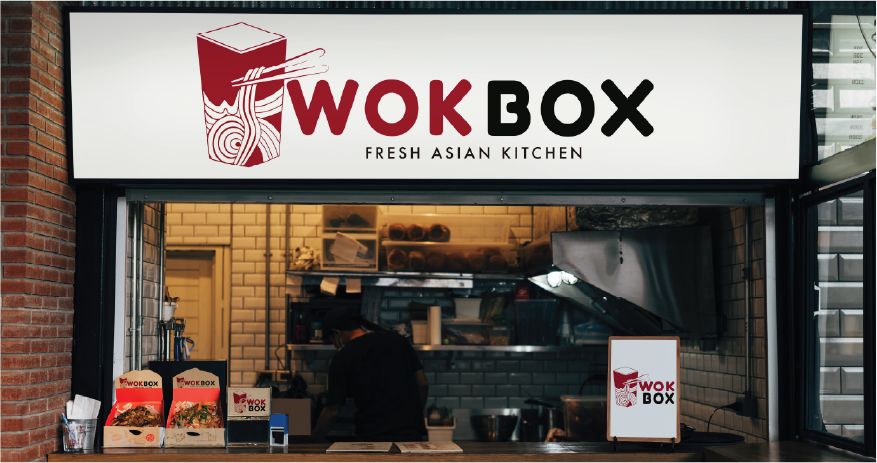 WokBox restaurant counter with illuminated sign showing red and black logo and slogan Fresh Asian Kitchen, with packaged food displayed and worker inside kitchen.