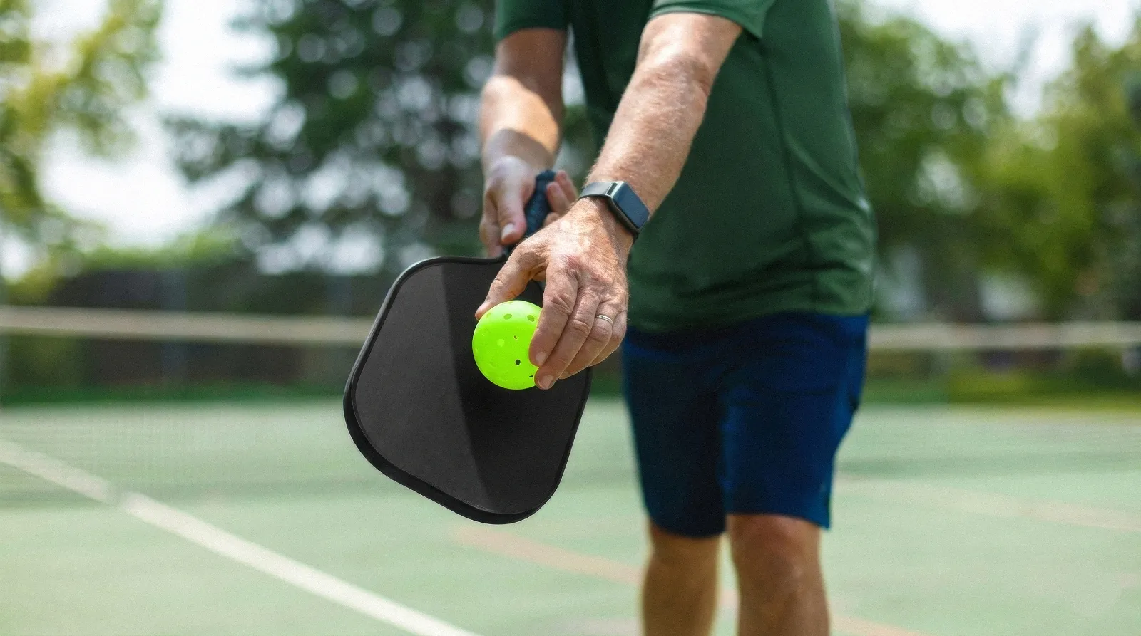 Person holding a pickleball paddle and bright yellow pickleball on an outdoor court.