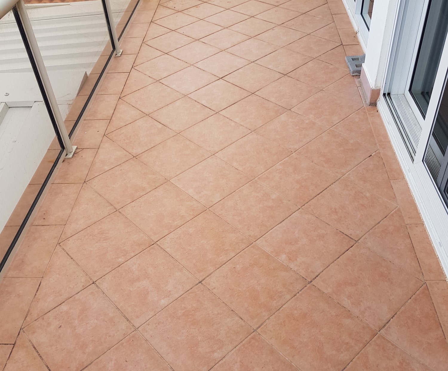Balcony floor with light brown square tiles arranged diagonally, glass railing on the left and sliding glass door on the right.