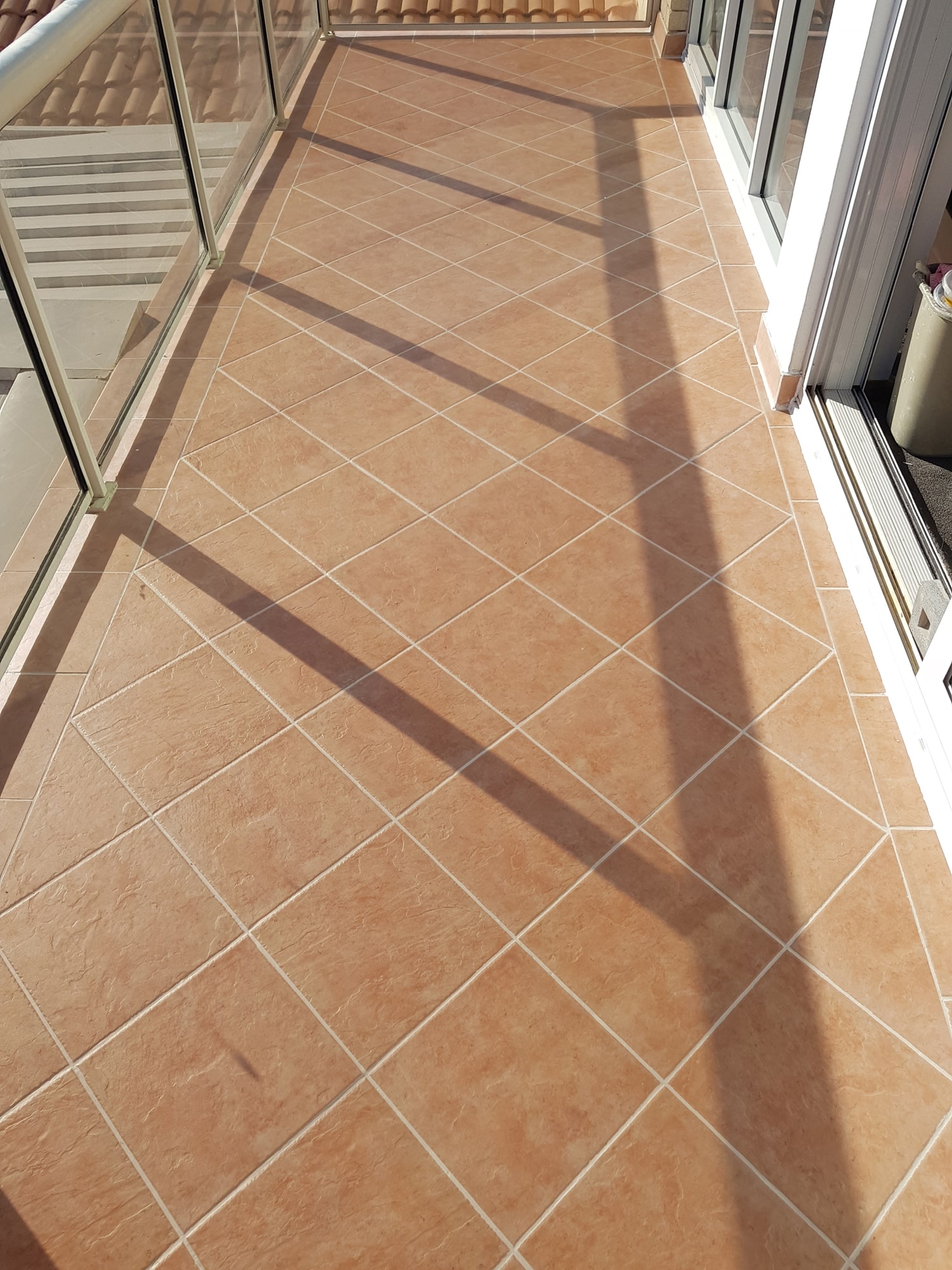 Sunlit balcony floor with beige tiles arranged in a diamond pattern and glass railing casting shadows.